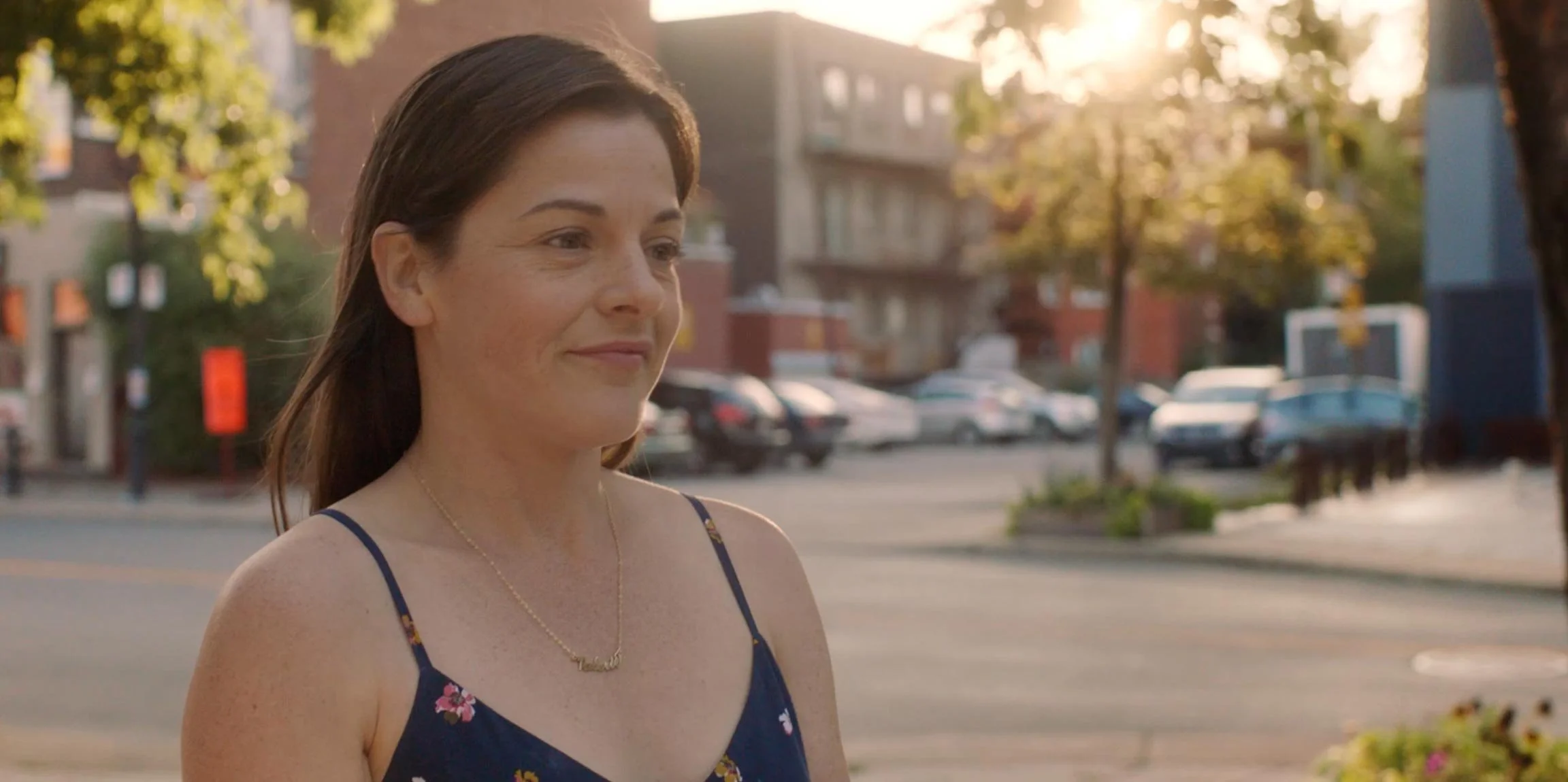 A woman with dark brown hair and fair skin standing outdoors during sunset, wearing a navy blue dress with floral patterns and a necklace, with an urban street and parked cars in the background.