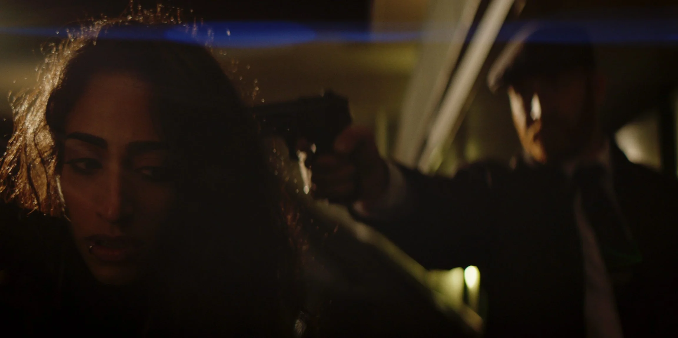 A woman with wavy hair looking to the side, partially illuminated with warm lighting, with a person in the background holding a gun to her head inside a vehicle at night.