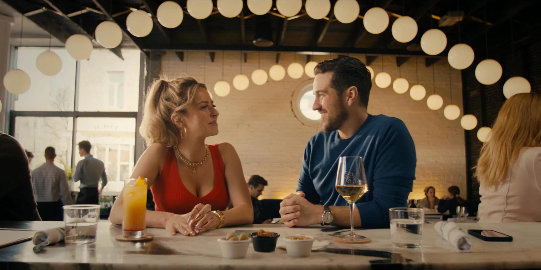A man and woman sitting at a restaurant table, smiling at each other, with drinks and small dishes on the table, inside a cozy, well-lit restaurant with hanging round lights.