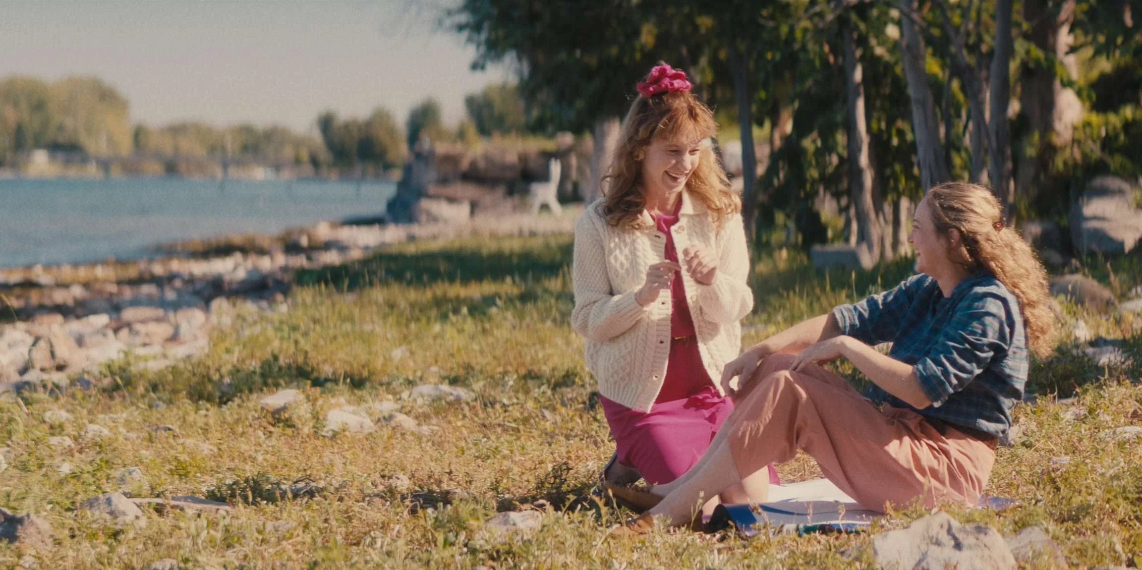 Two women with curly hair are on the ground near a body of water, smiling and engaging in conversation on a sunny day, surrounded by trees and rocks.