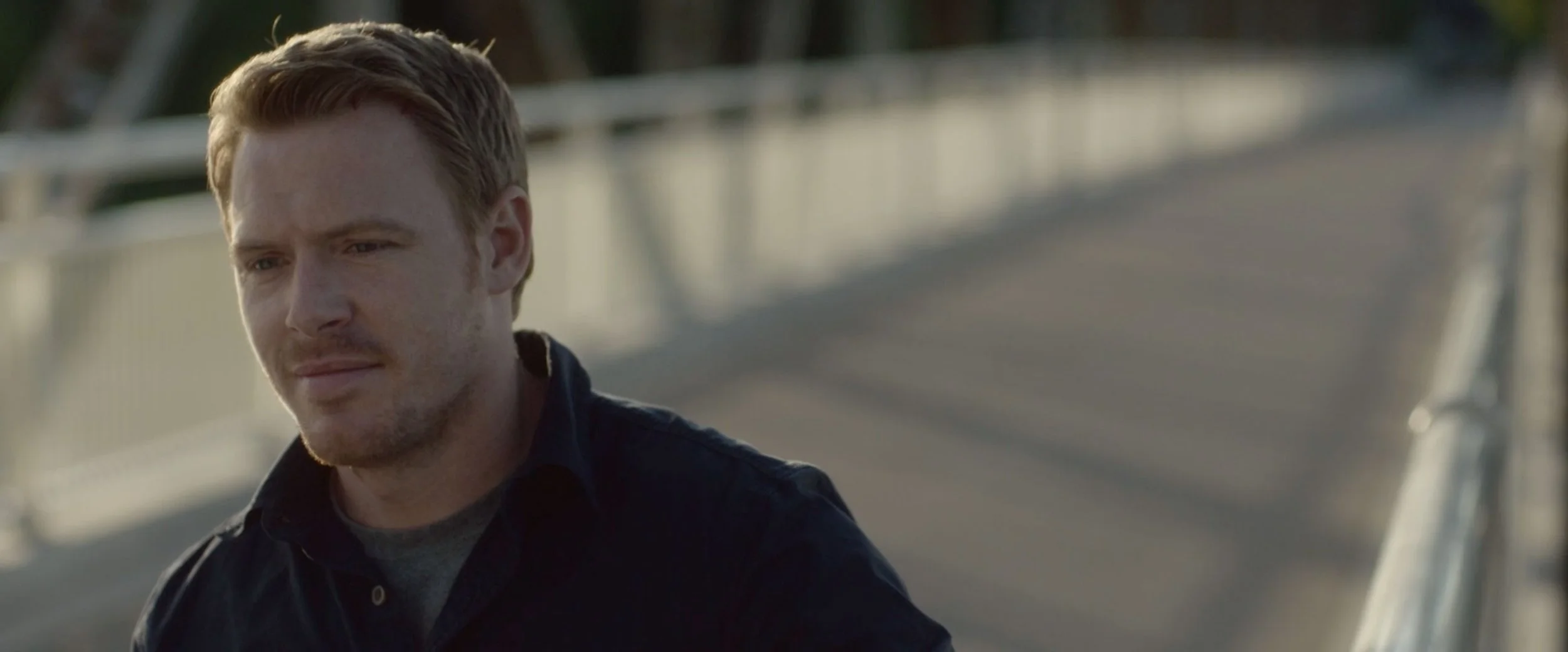 A young man with light brown hair and a slight beard, wearing a dark jacket and grey shirt, looking thoughtful while sitting on a bridge with a metal railing.