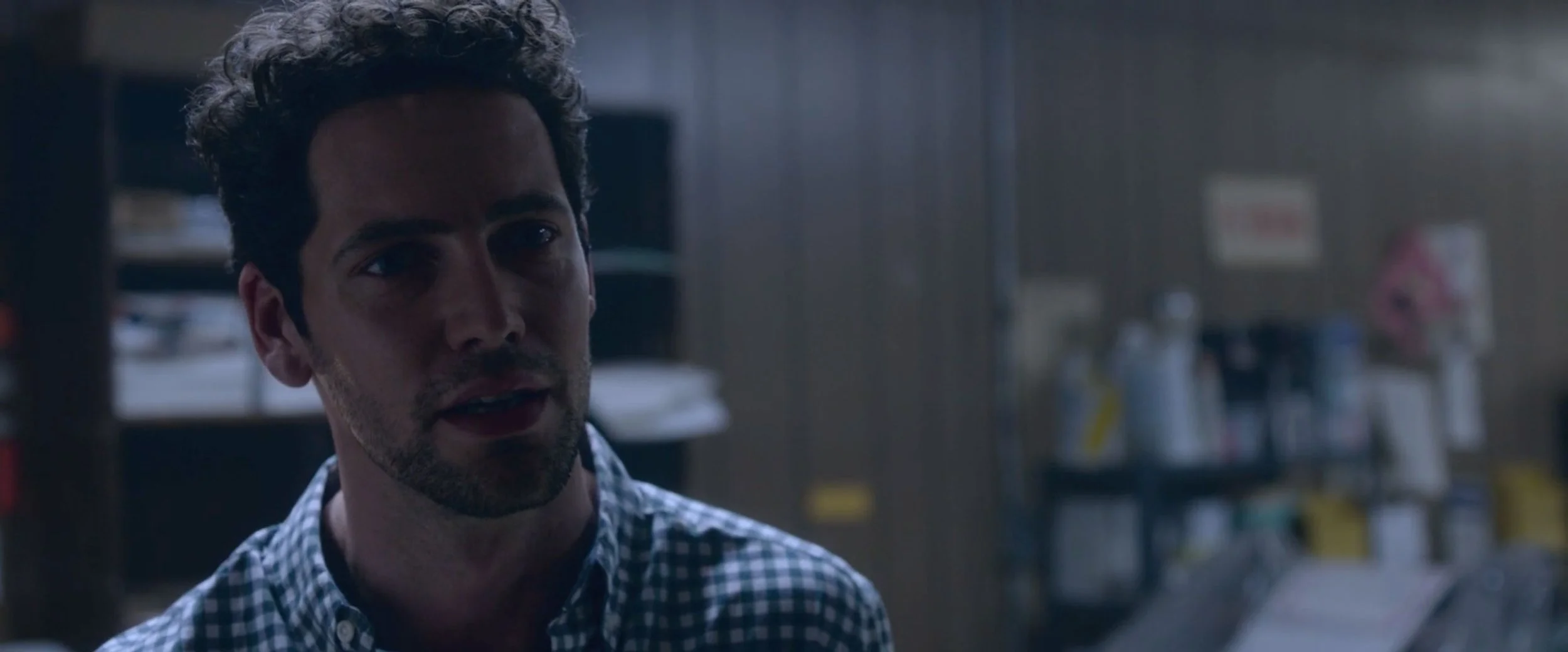 A man with dark hair and a beard, wearing a checkered shirt, standing in what appears to be a store or office with shelves and various items in the background.