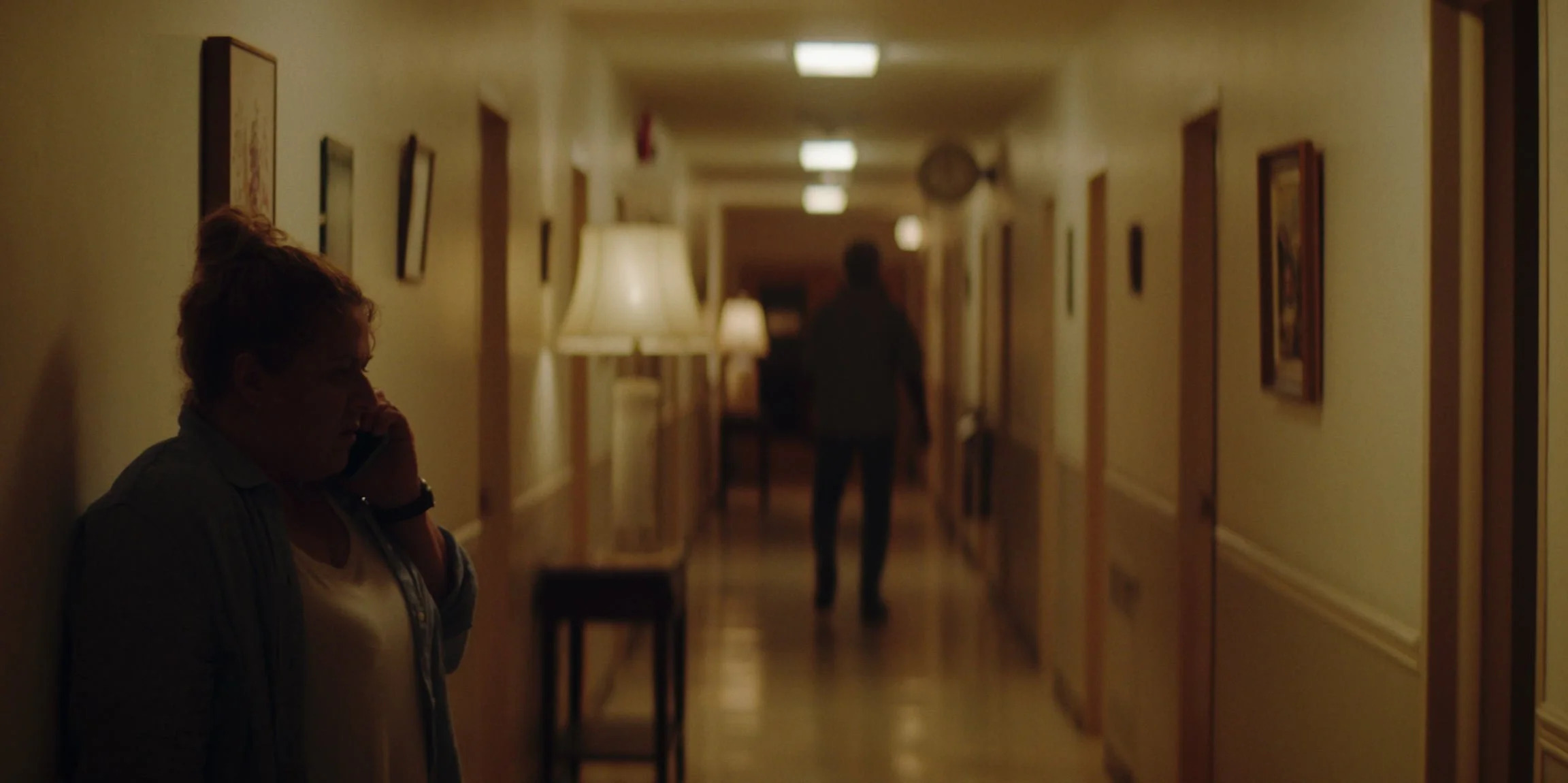 A woman with red hair talking on the phone while leaning against a wall in a dimly lit hotel hallway, with another person walking away in the background.