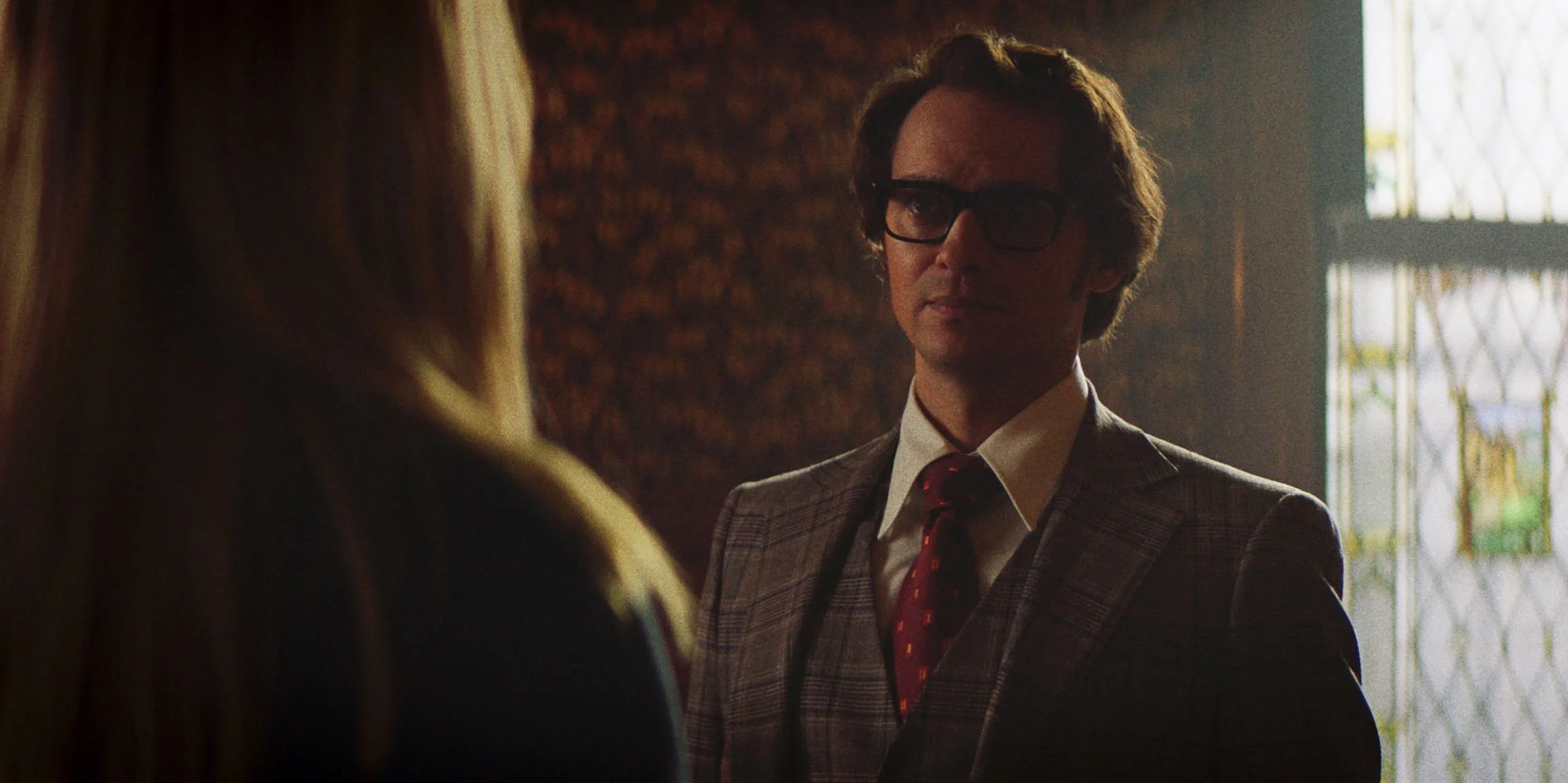 A man in a plaid suit and tie, wearing glasses, converses with a woman inside a room with stained glass windows, soft lighting, and textured walls.