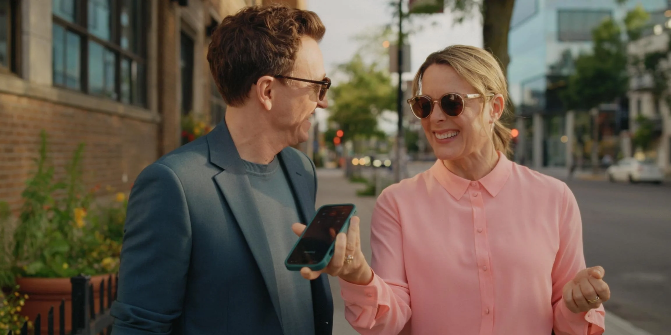 A man in a blue blazer and a woman in a pink blouse are smiling and talking while walking outdoors on a city street during daytime.