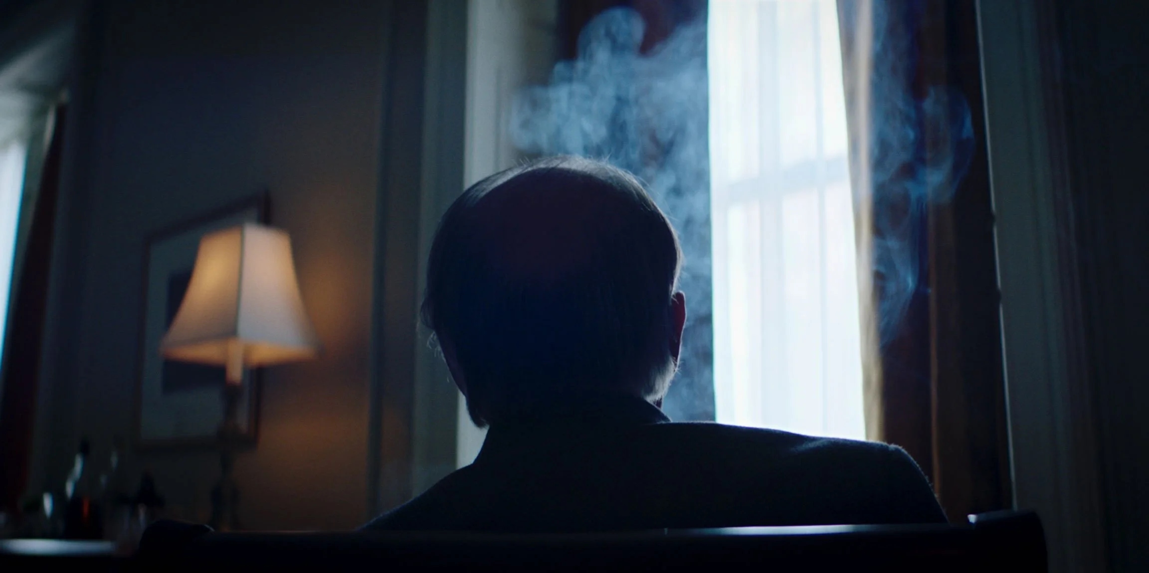 Person sitting in a dimly lit room, facing a window with curtains, with visible smoke or vapor around them.