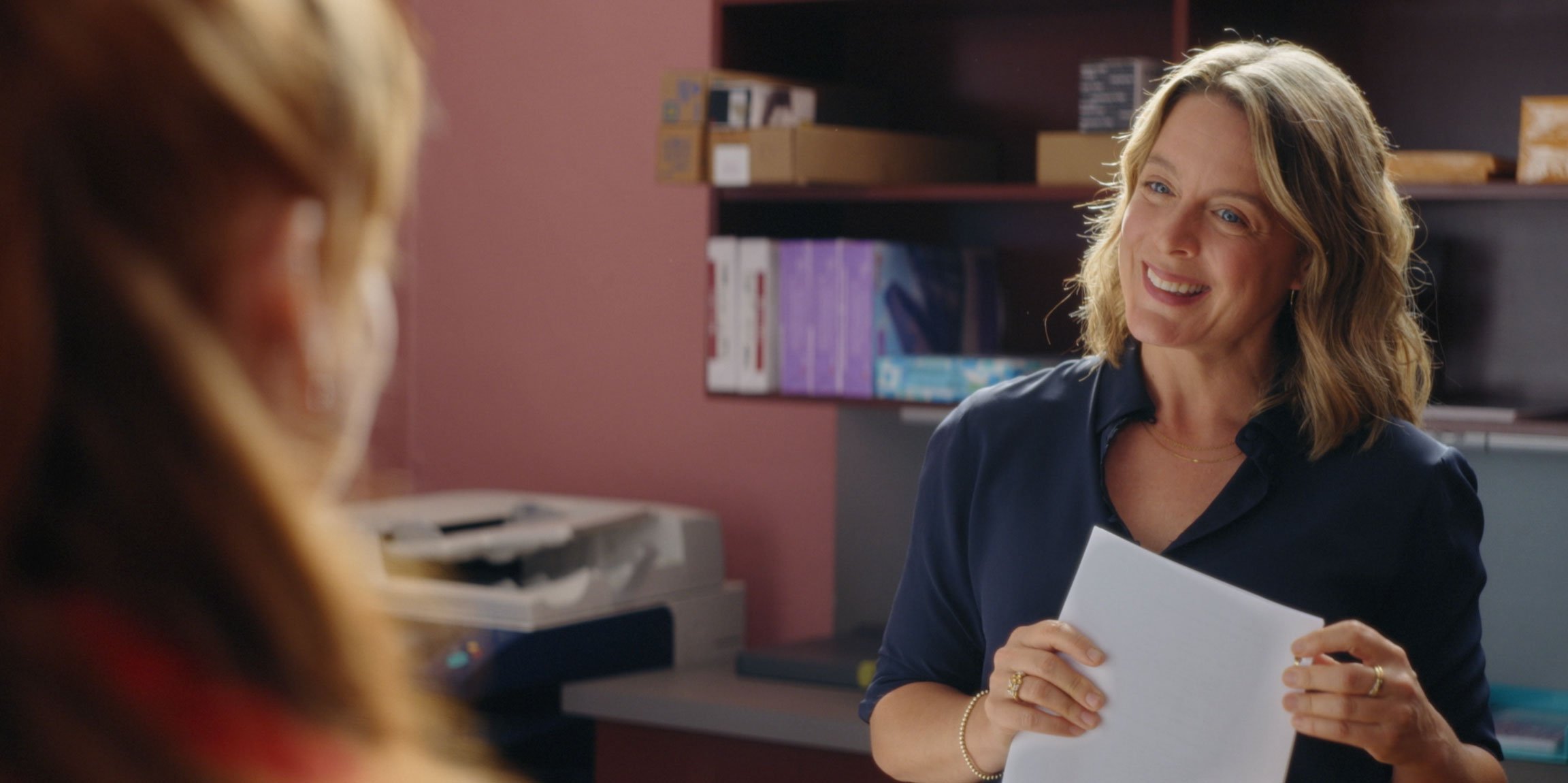 A woman in a navy blue shirt smiling and holding white papers, engaged in conversation with another person, in an office setting.
