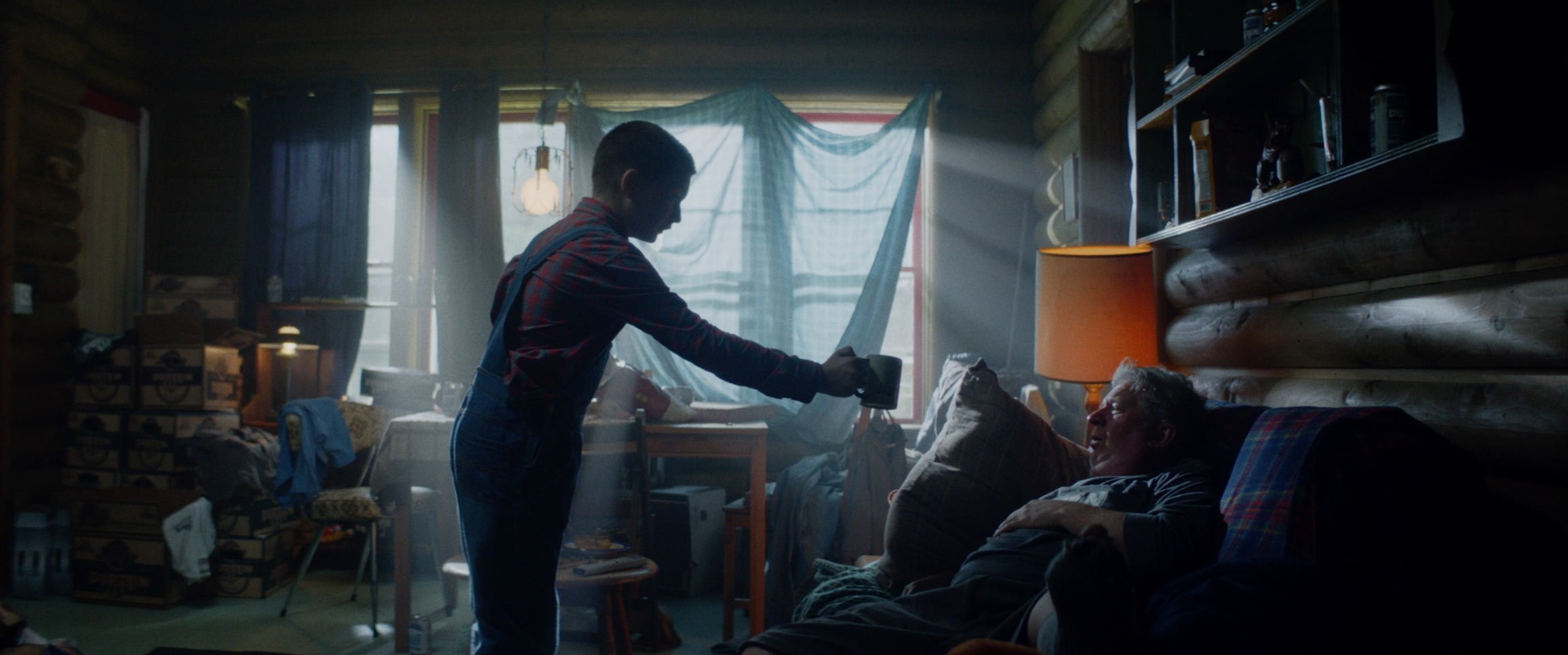 A young man with a coffee pot offers coffee to an elderly man lying in bed inside a cozy room with wooden log walls, a lamp, and cluttered furniture. Light streams through a window covered with fabric and curtains.