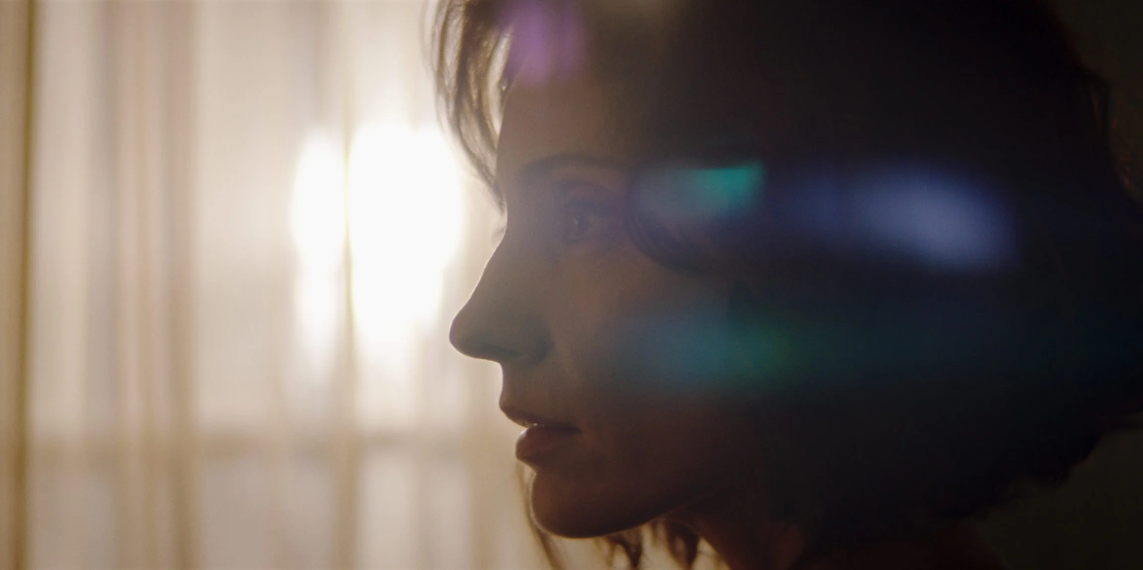 Close-up side profile of a woman with her eyes closed, backlit by sunlight coming through a window, with lens flare effects.