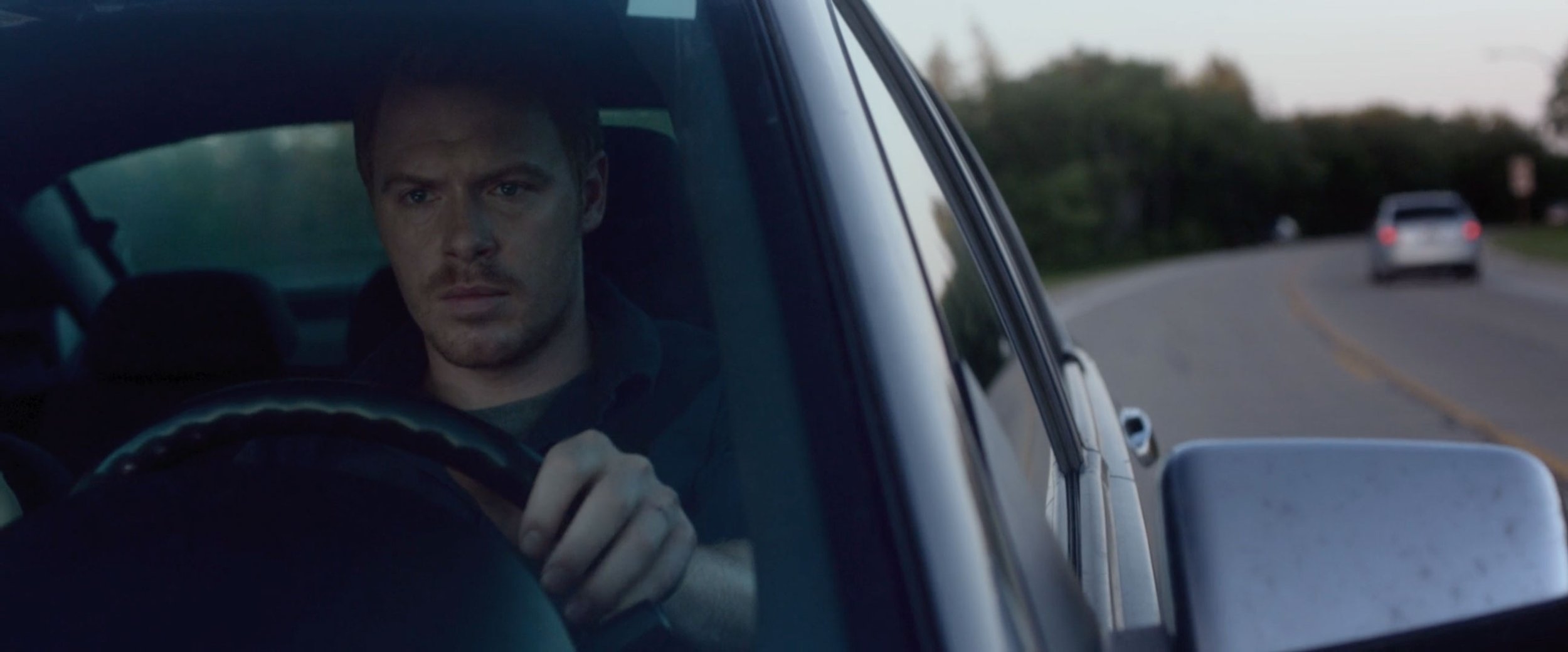 A man with a serious expression driving a car on a winding road during dusk or dawn