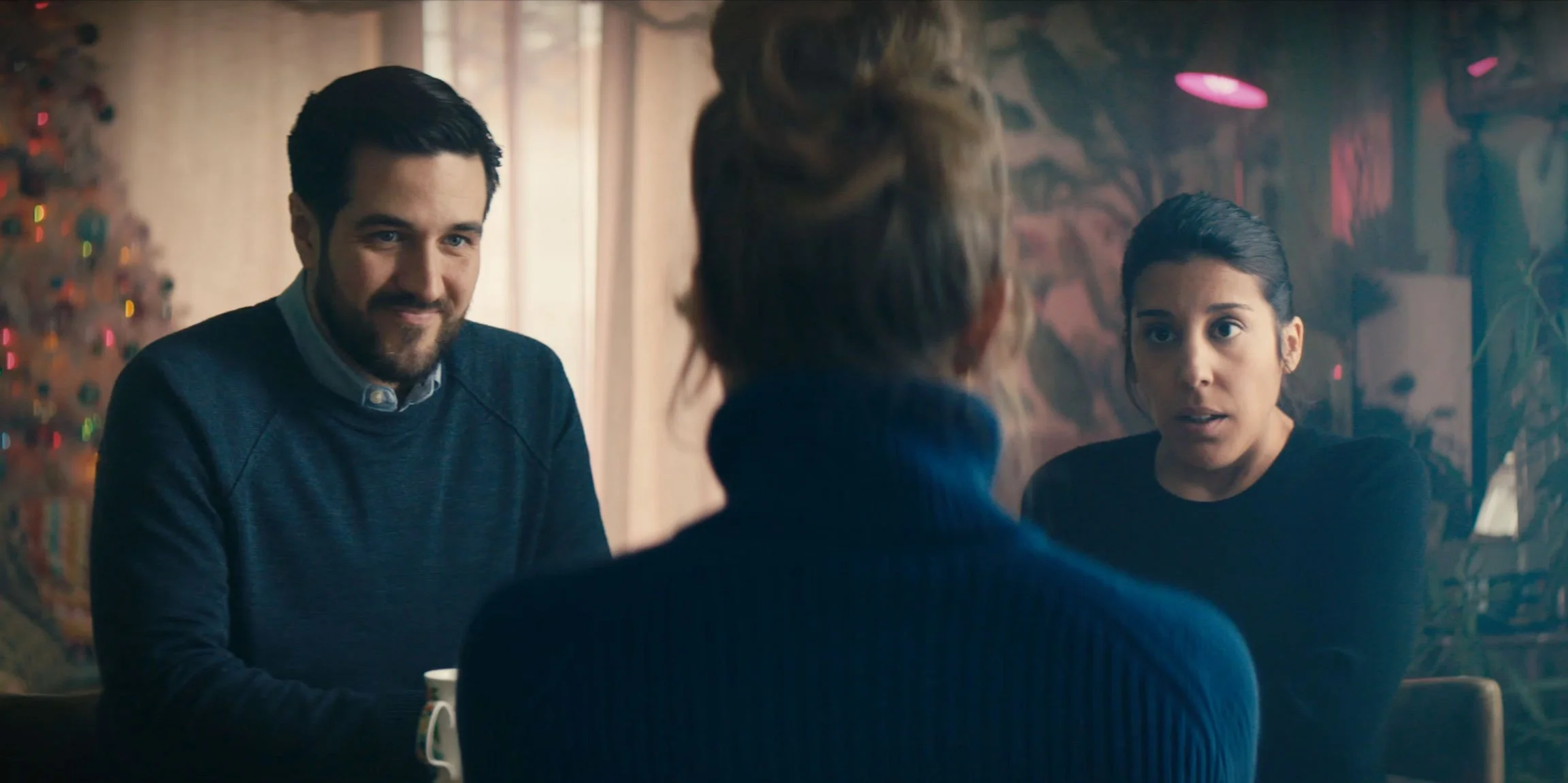 Three people having a conversation at a table, one man smirking, two women listening attentively in a cozy, decorated room.