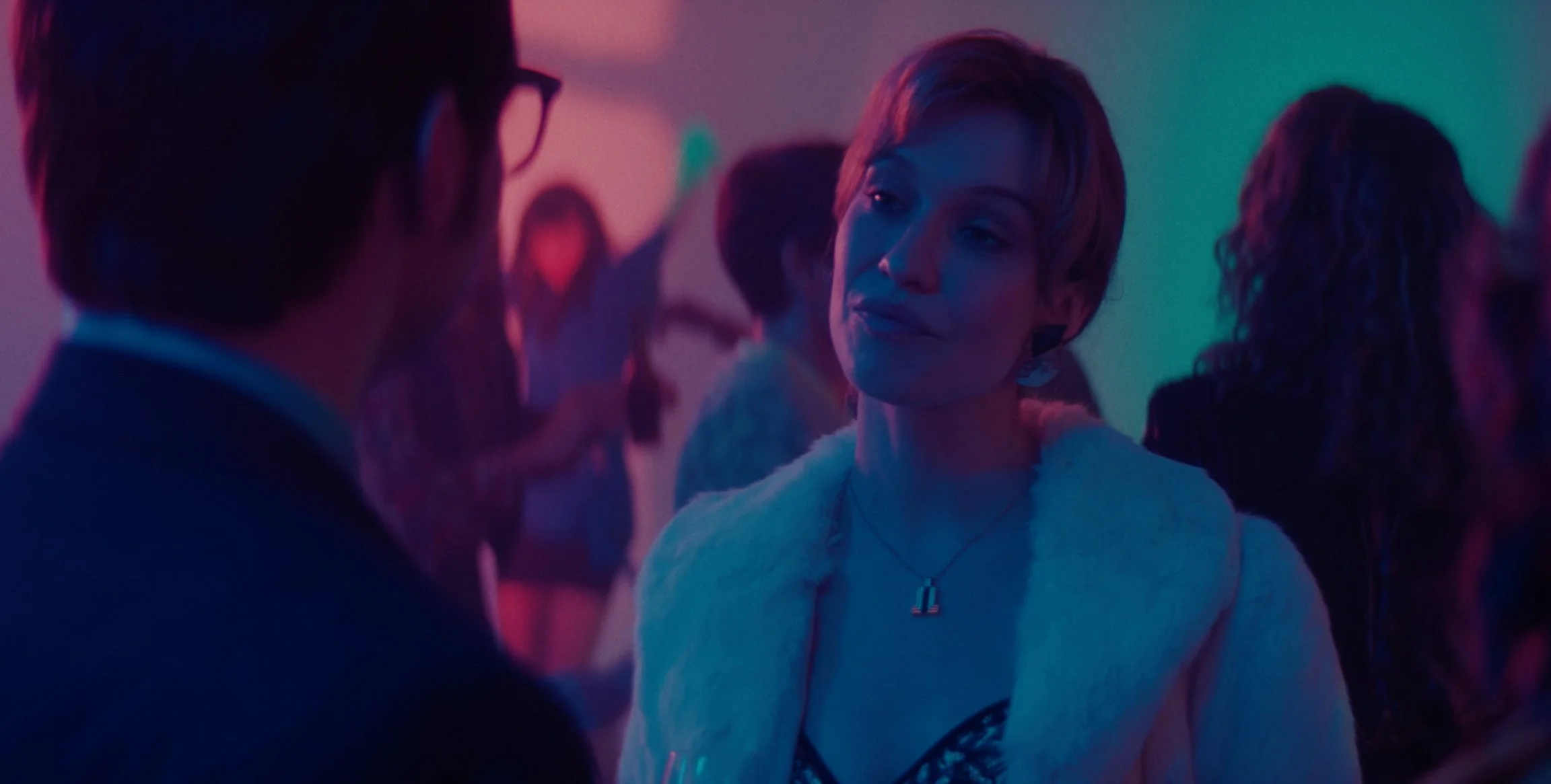 A woman with short hair and a white fur coat talking with a man in glasses in a dimly lit party with colorful lighting.