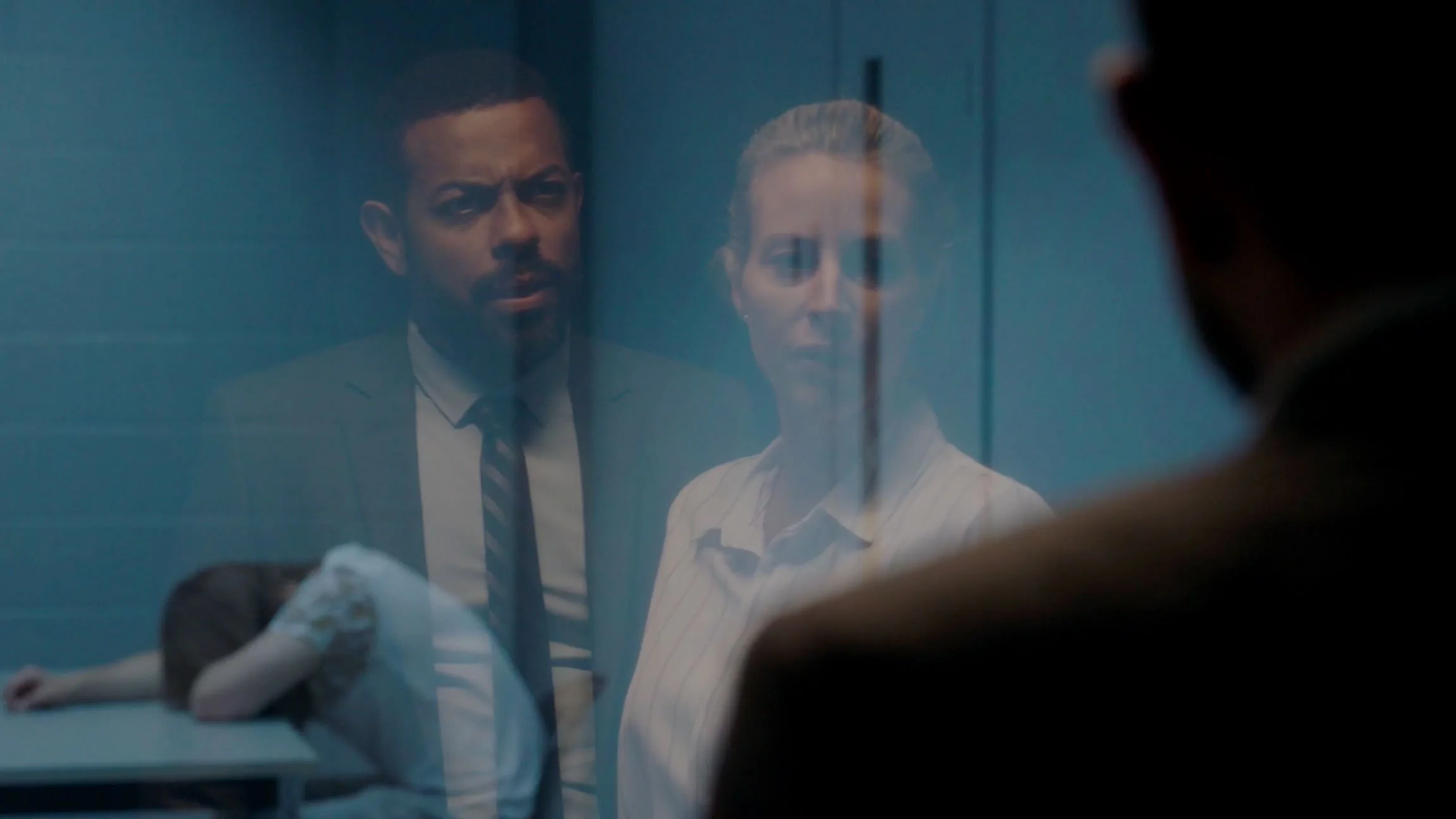 Two people behind a glass window, a woman stands and looks serious, a man looks confused, with a person lying motionless on a table in the foreground.