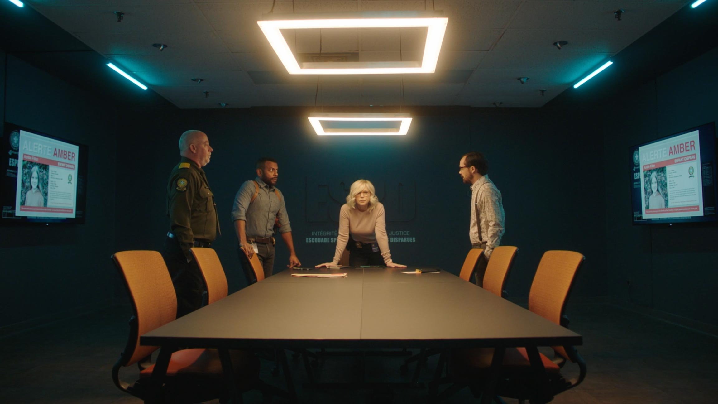 Four people standing around a conference table in a dark room with two monitors displaying an alert about someone named Amber.