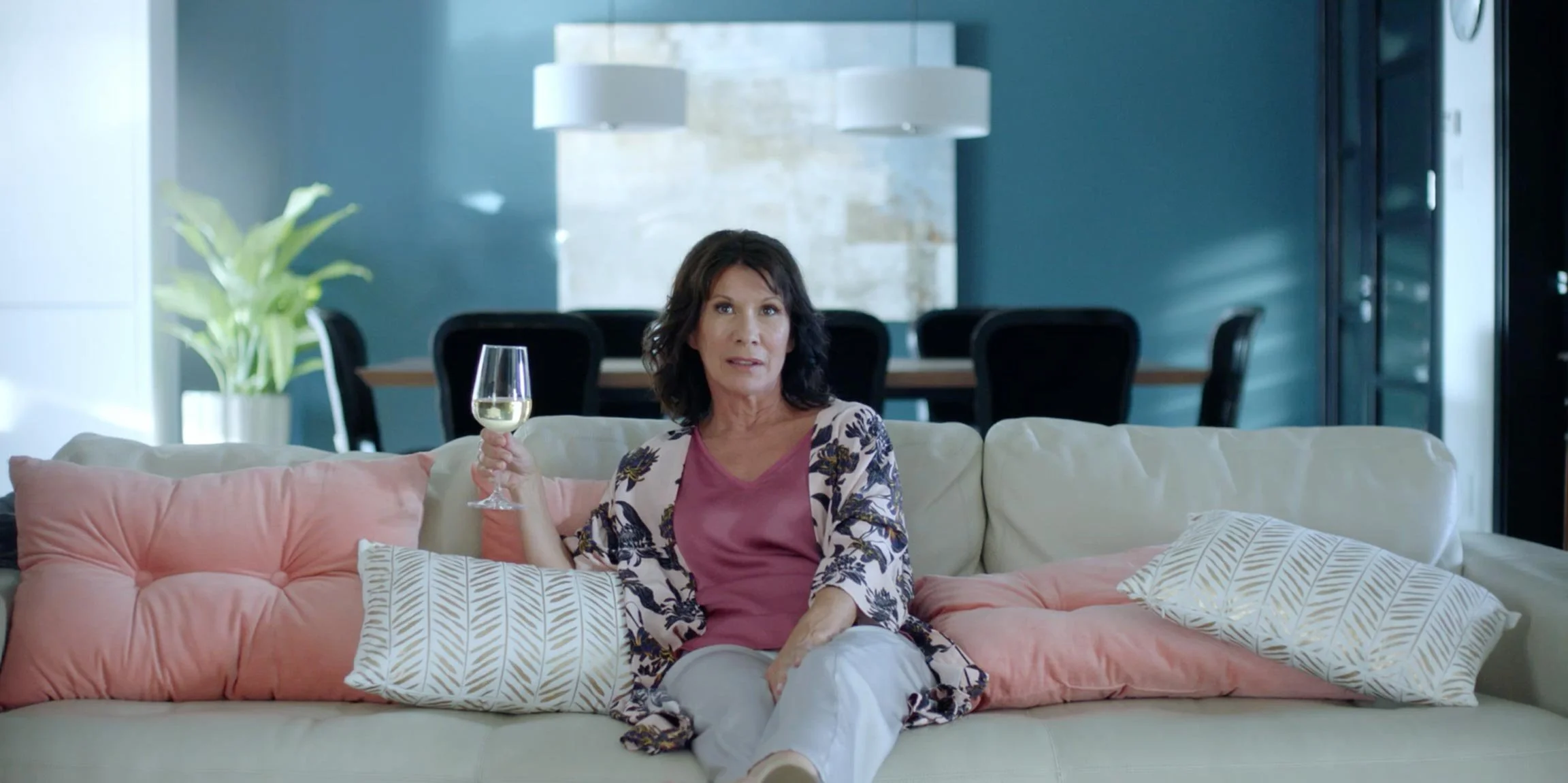 A woman sitting on a beige sofa in a modern living room, holding a glass of white wine, with decorative pillows and a potted plant in the background.