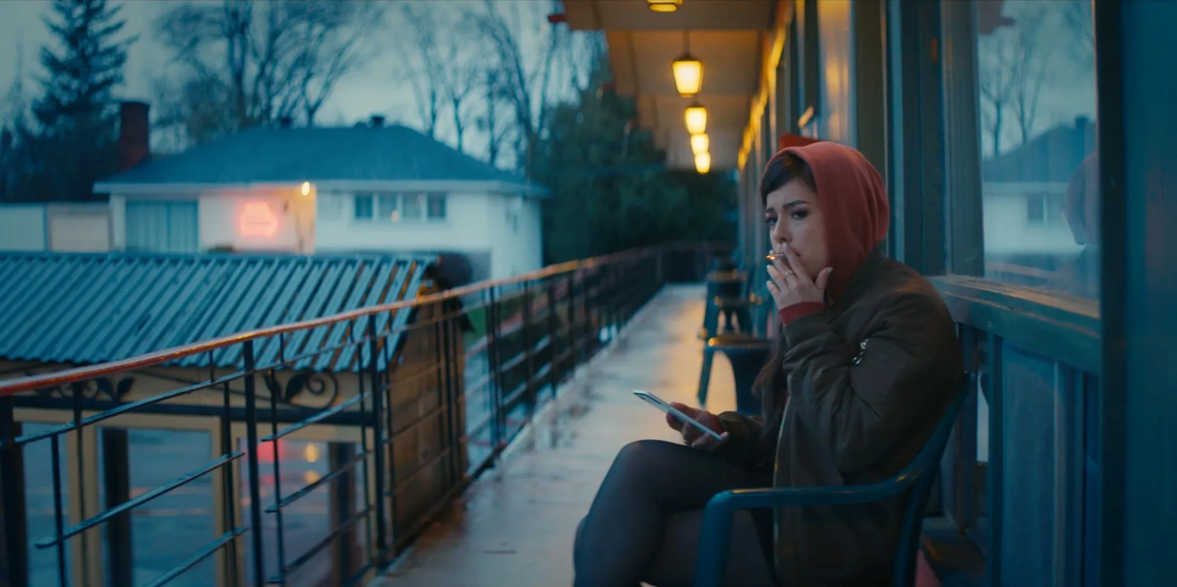 A young woman with short dark hair wearing a red hoodie and a brown jacket sitting on a bench at a dimly lit outdoor train station platform, smoking a cigarette, and looking at her phone with a contemplative expression. The background features a hous