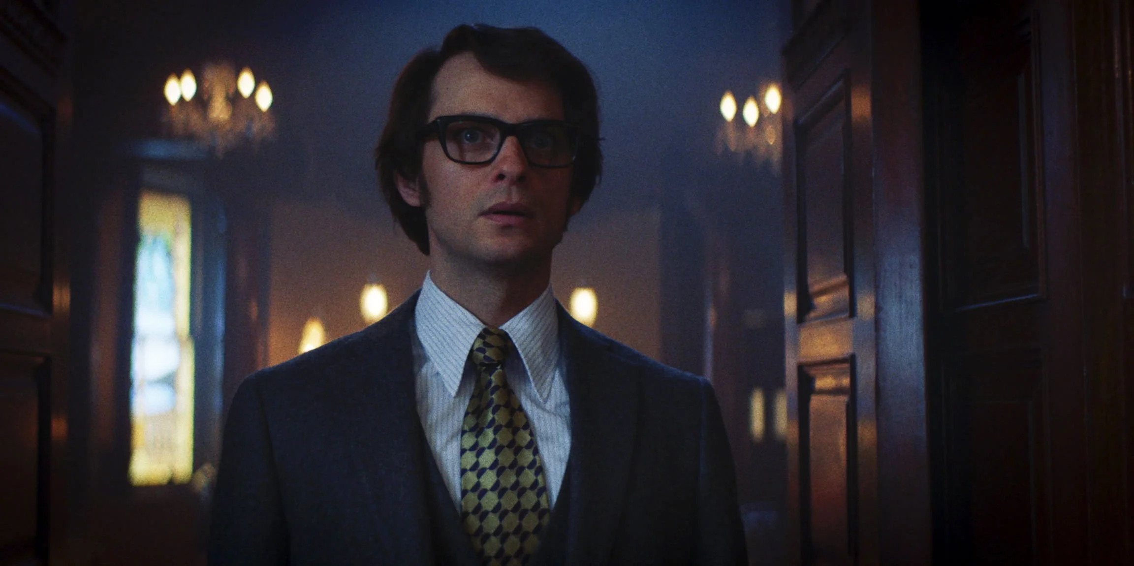 A man with dark brown hair and glasses wearing a dark jacket, white shirt, and patterned tie, standing in a dimly lit room with wooden paneling and stained glass windows.