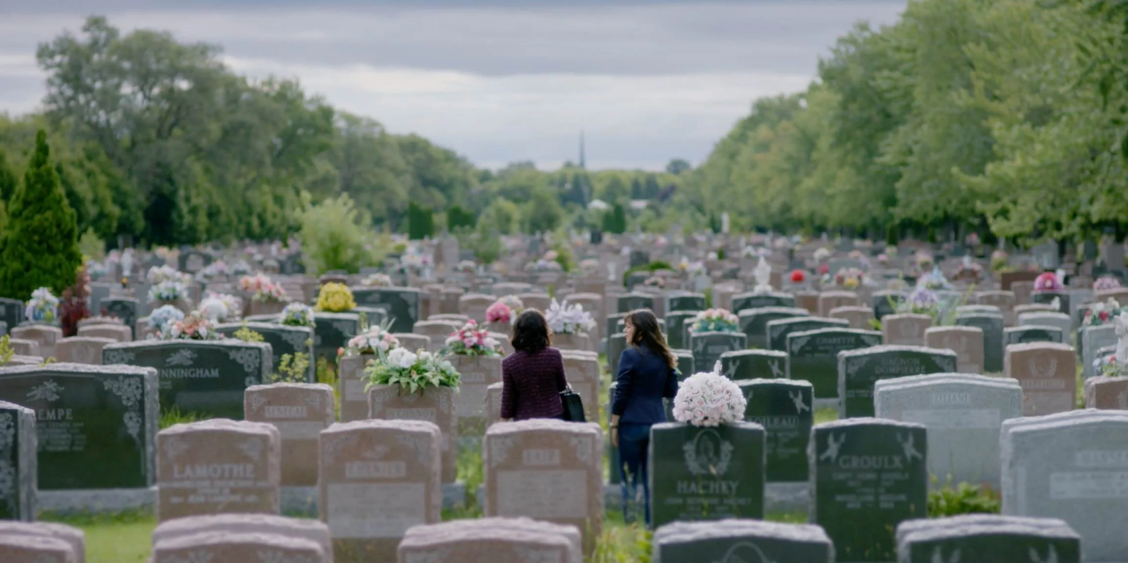 Two women walking and talking through a cemetery filled with gravestones decorated with flowers, under cloudy skies.