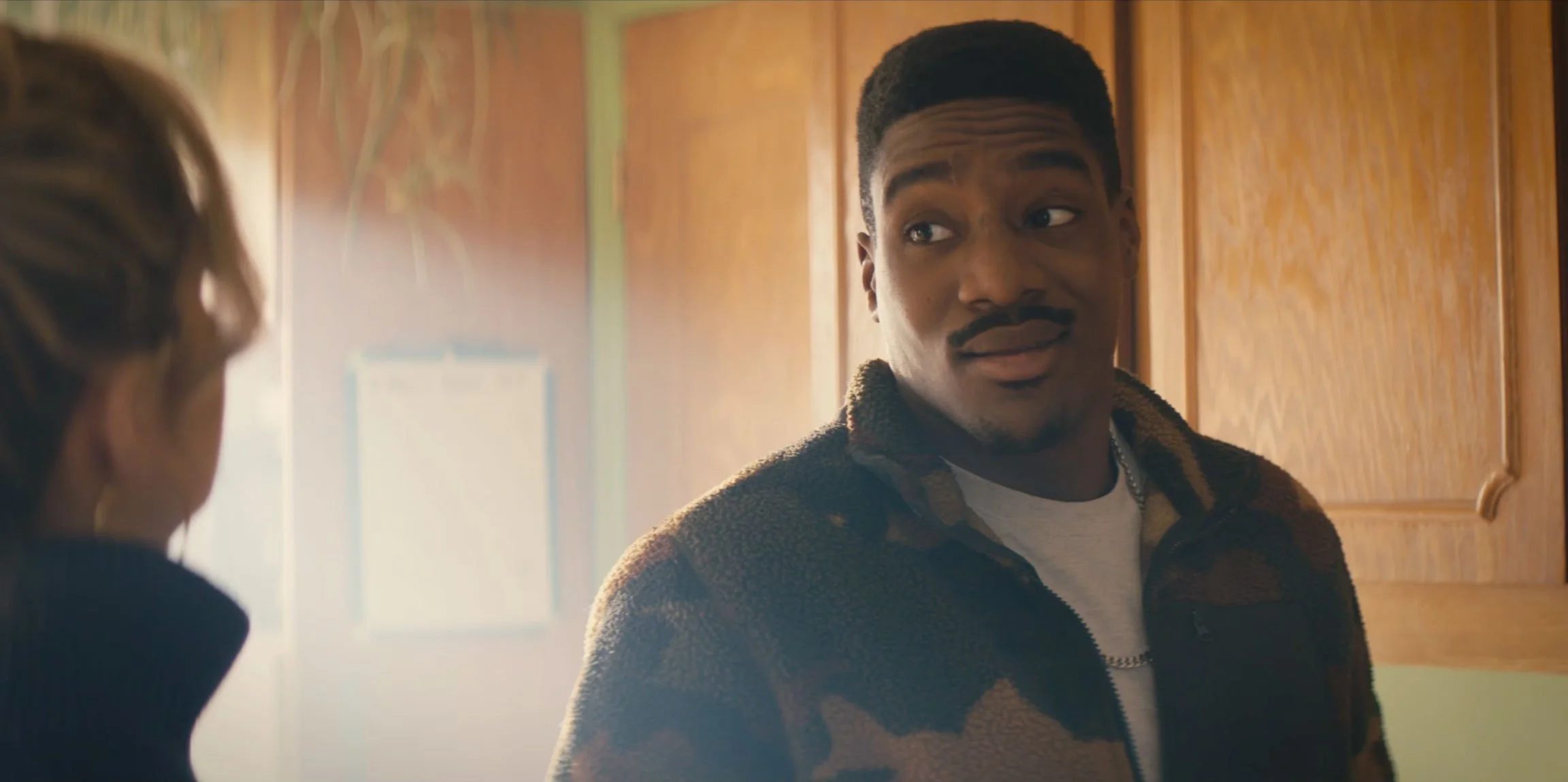 A young man with short dark hair and a mustache looking at a woman in a kitchen with wooden cabinets, wearing a patterned fleece jacket.