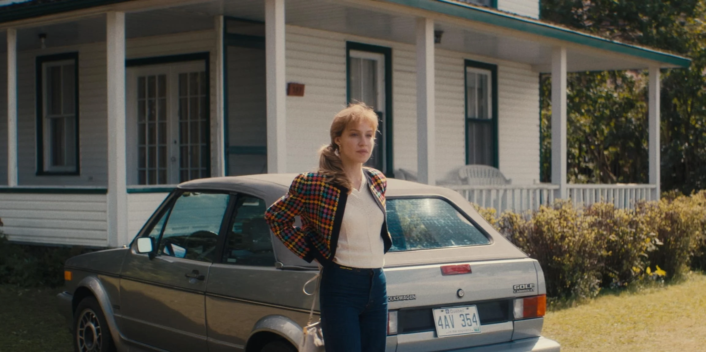 A woman stands next to a vintage gray Volkswagen Golf car in front of a white house with a porch, green trees in the background, and sunlight illuminating the scene.