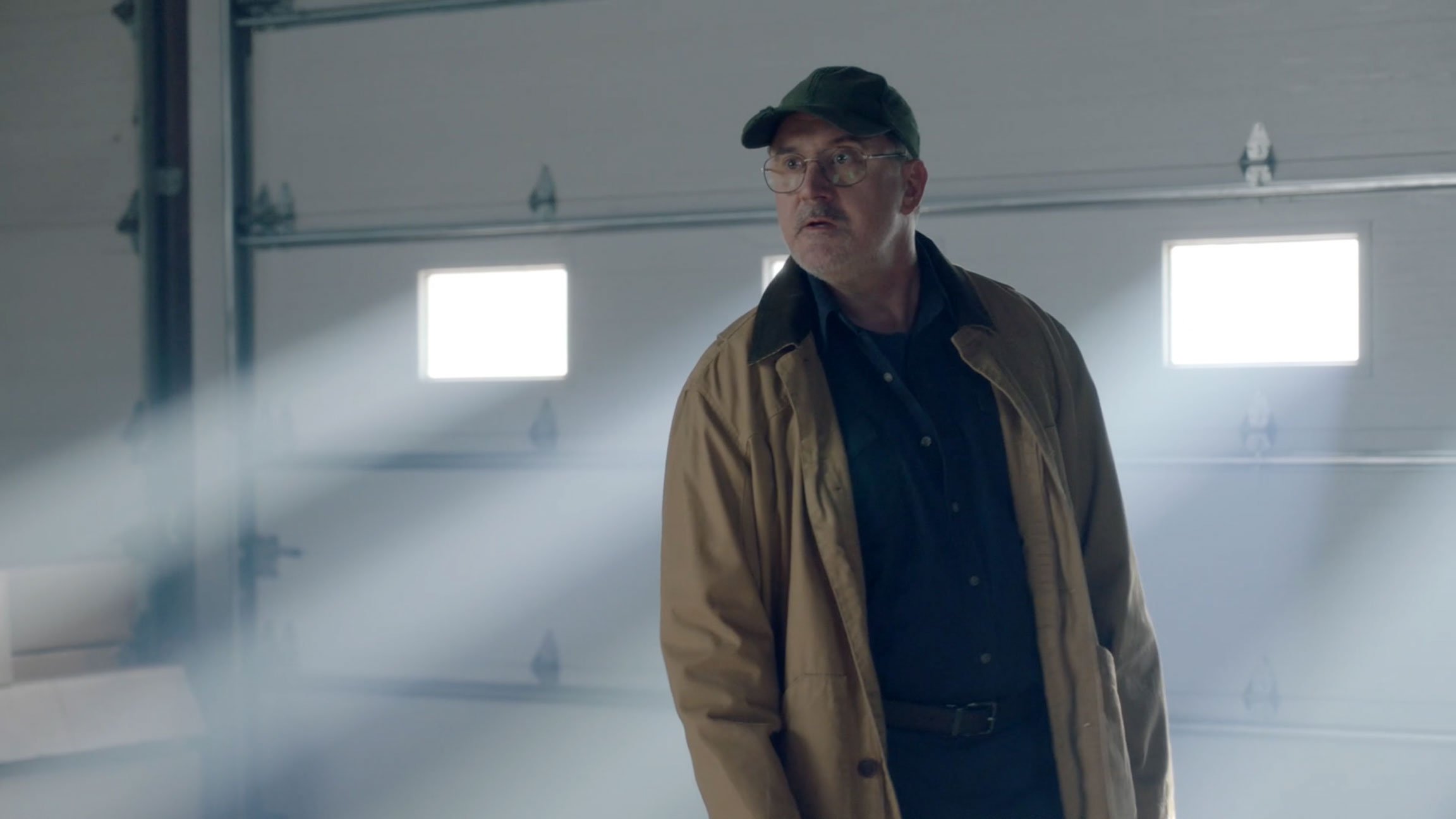 A middle-aged man wearing glasses, a cap, and a tan jacket standing in a garage with a large white door, illuminated by small windows.