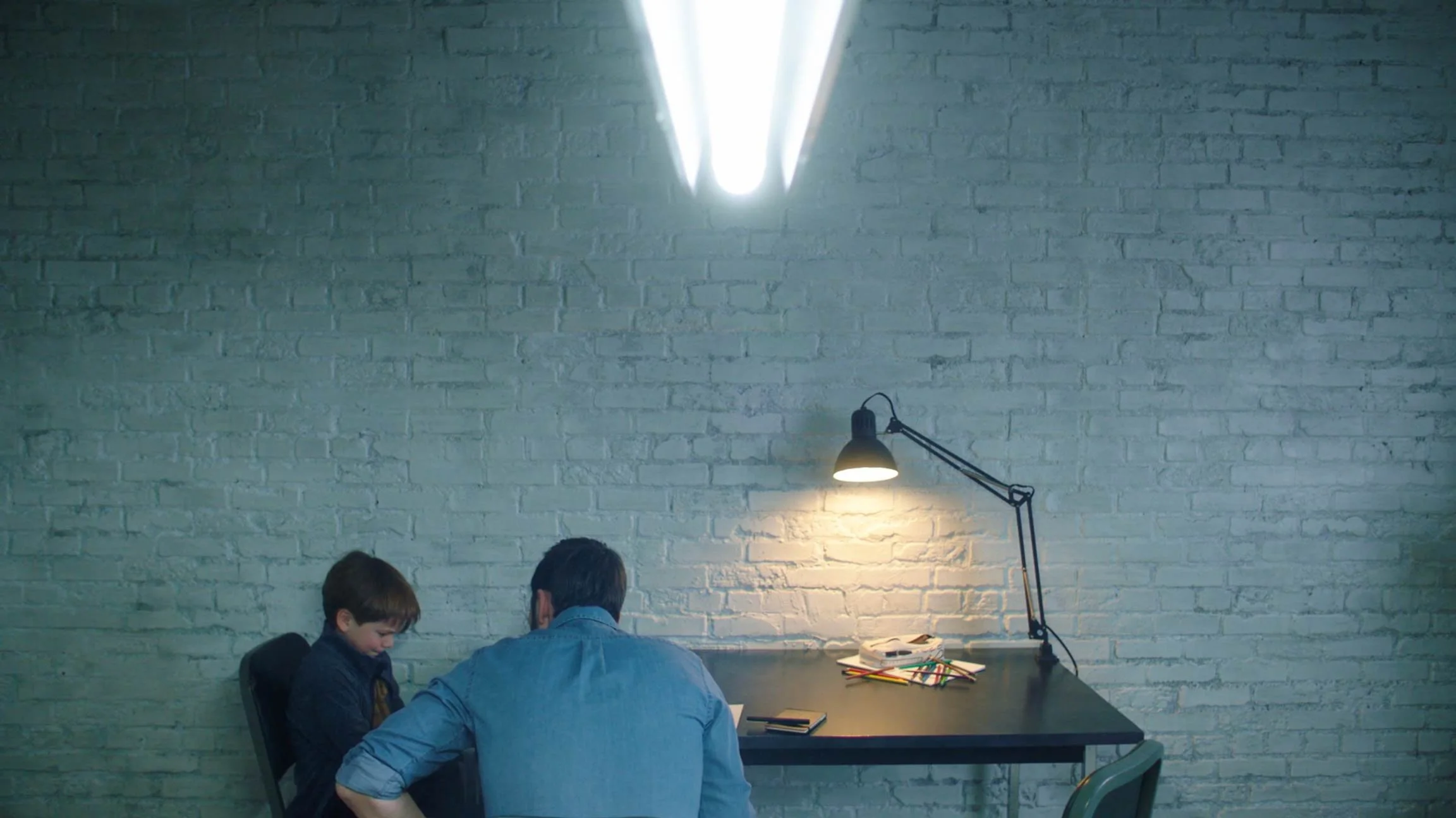 A man and a boy sitting at a desk in a room with white brick walls, illuminated by a desk lamp and an overhead fluorescent light.