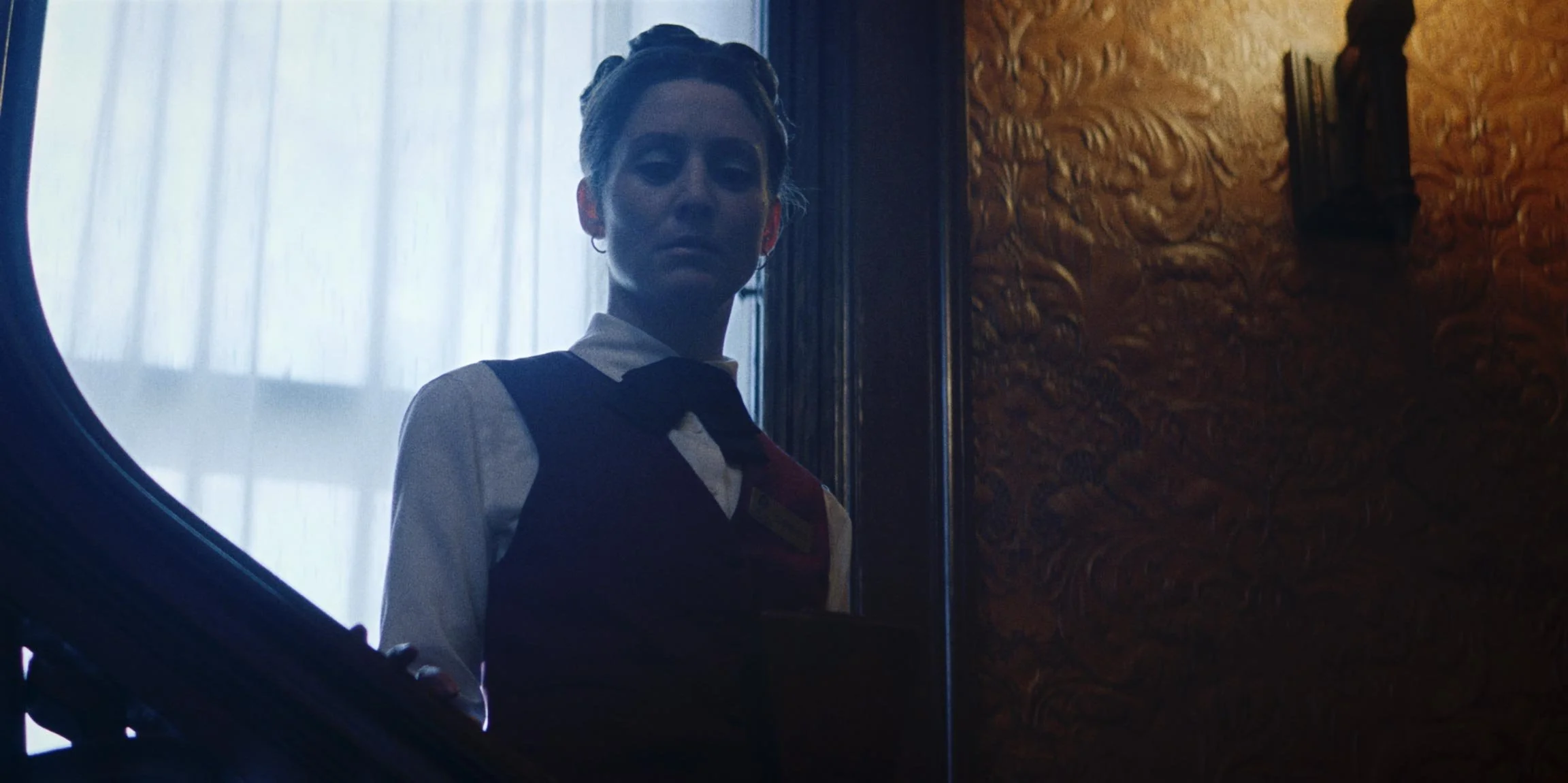 Woman in a uniform standing in front of a mirror with curtains and ornate wood paneling behind her.