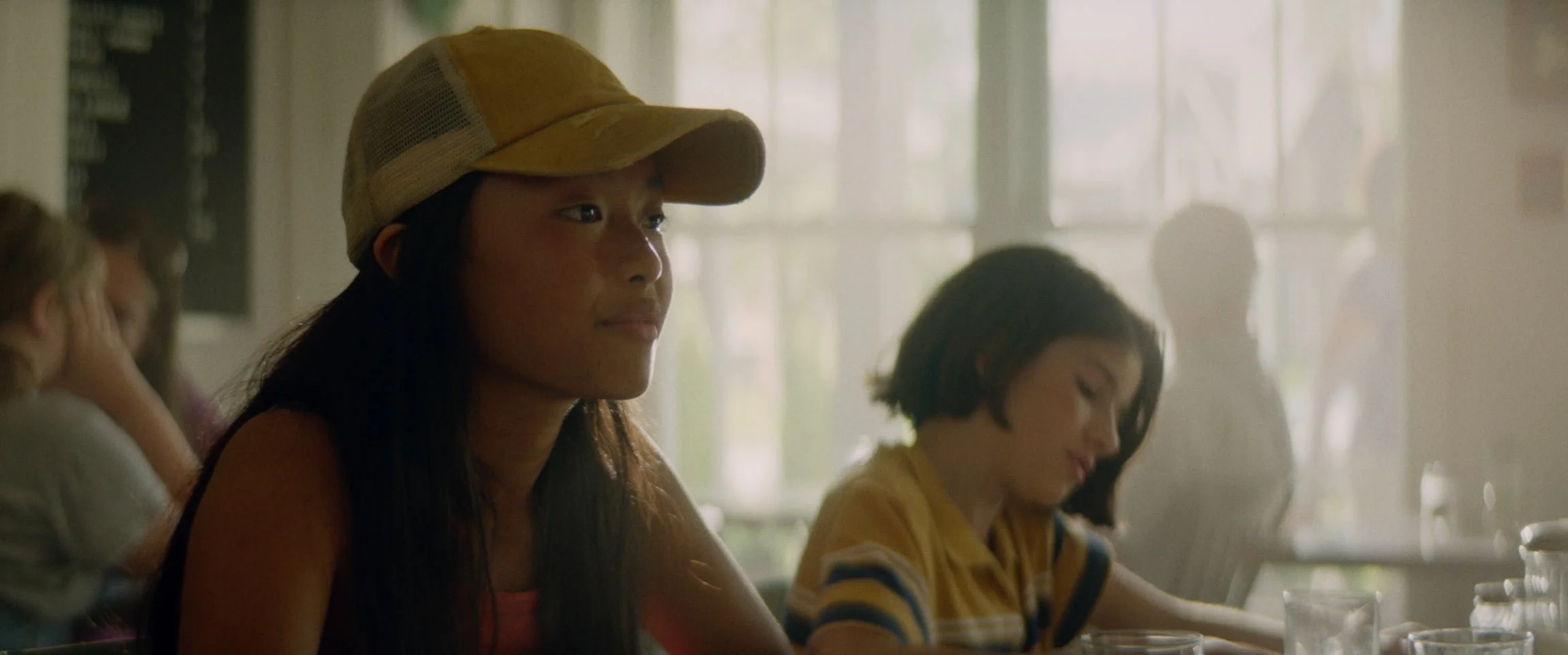 Young woman with long dark hair wearing a yellow cap sitting at a table in a brightly lit room, with children sitting nearby.
