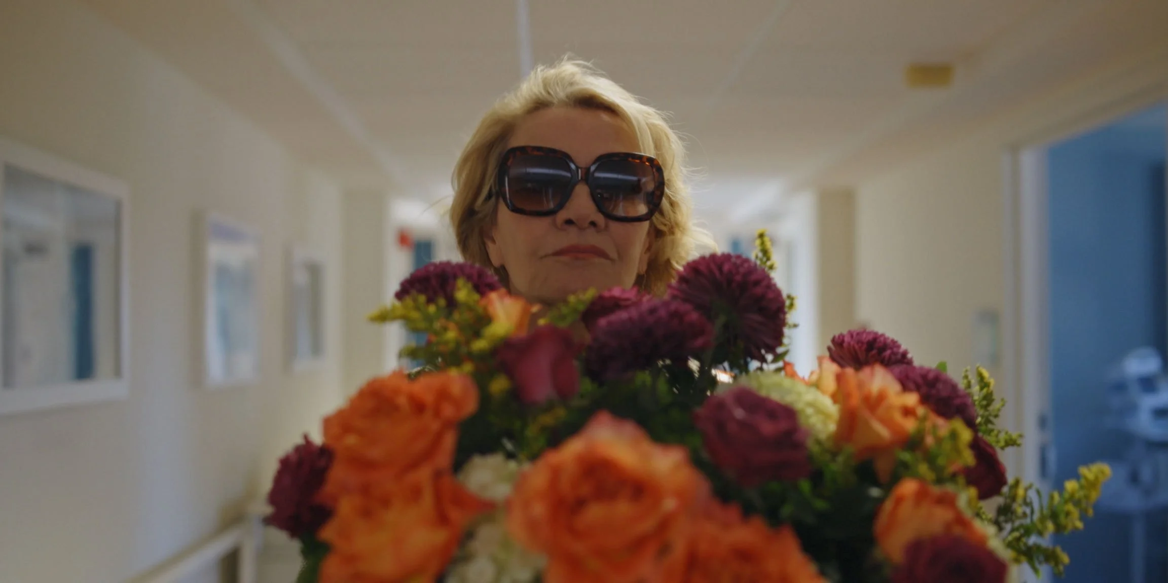 An older woman wearing large dark sunglasses holds a bouquet of colorful flowers in a hallway.