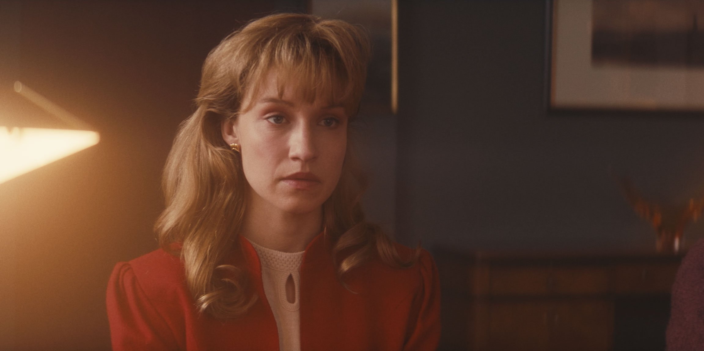 A young woman with shoulder-length wavy red hair and fair skin, wearing a red blazer and a white blouse, sitting indoors with a serious, contemplative expression.