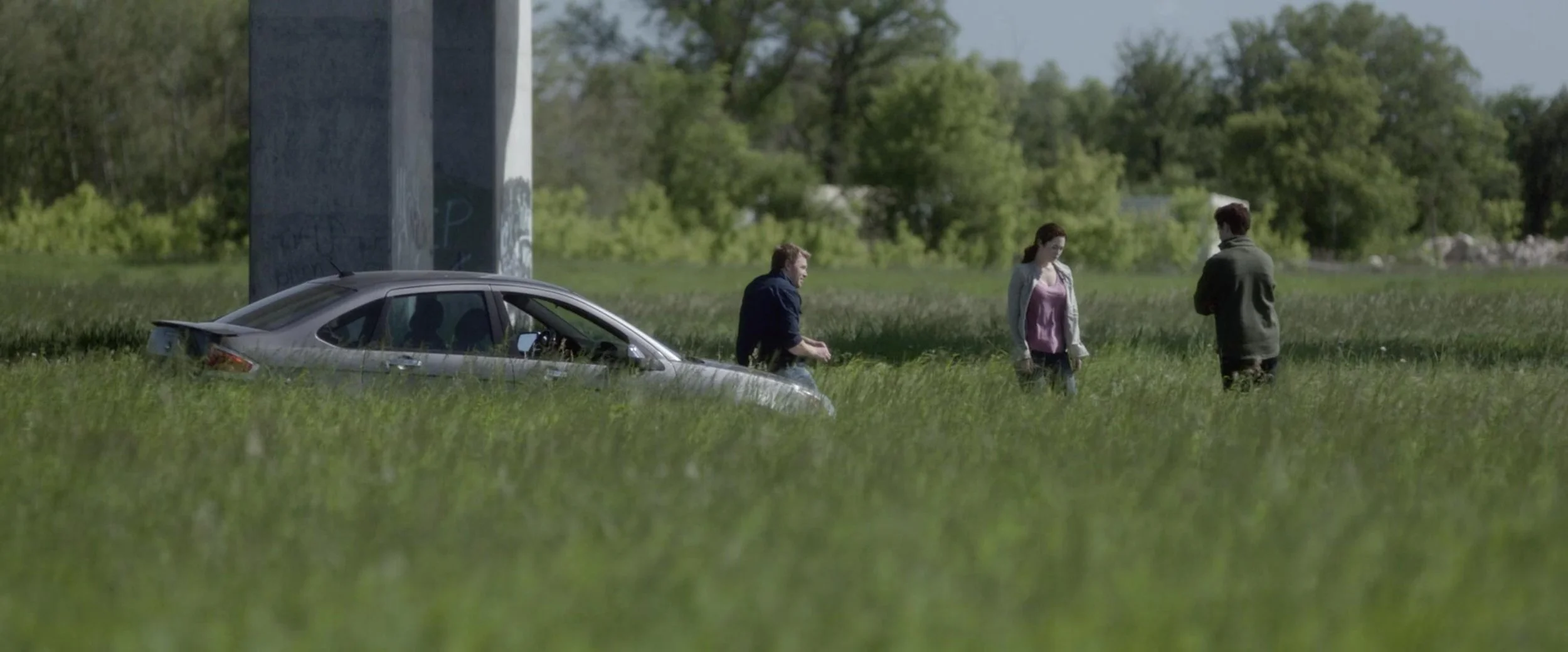 Three people standing in a grassy field near a silver car partially buried in the grass, with a large concrete pillar in the background and trees beyond.