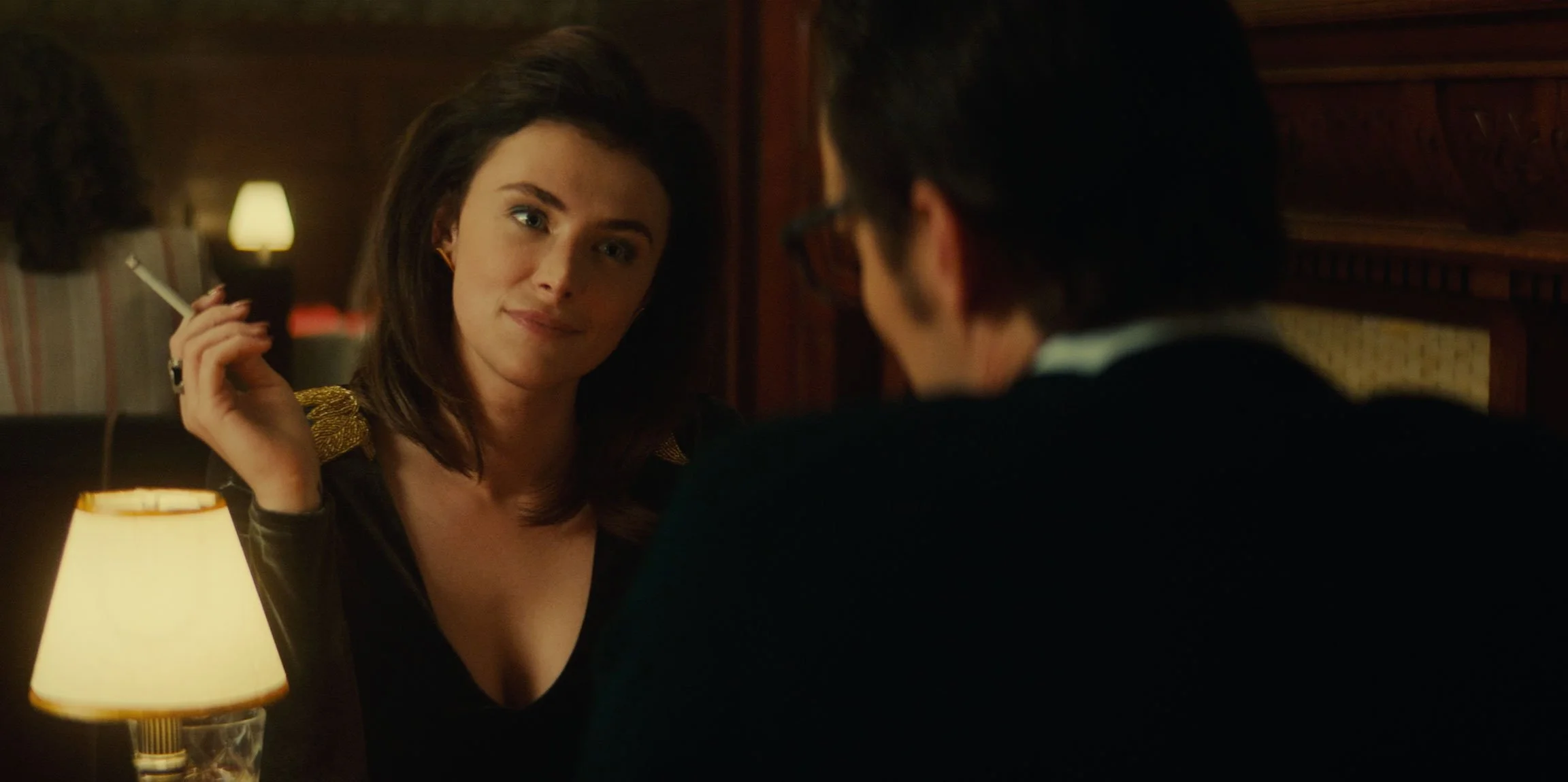 A woman with dark hair and wearing a black dress with gold accents, sitting at a dimly lit table, holds a cigarette in her right hand, and looks at a man with glasses, dark hair, and a black sweater. The scene appears to be from a movie or TV show se