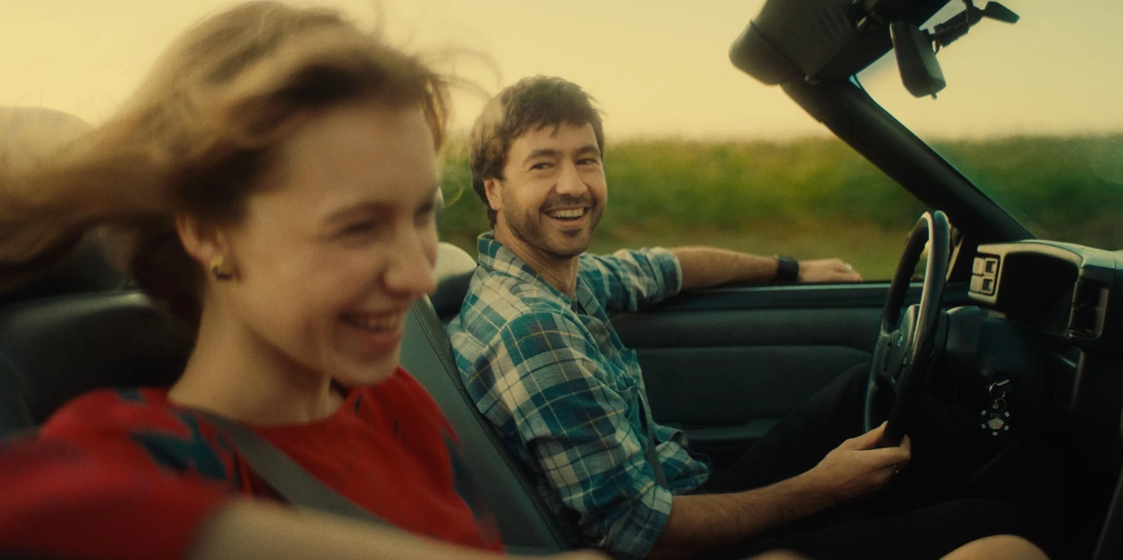 A smiling couple driving in a convertible car through a countryside landscape at sunset.