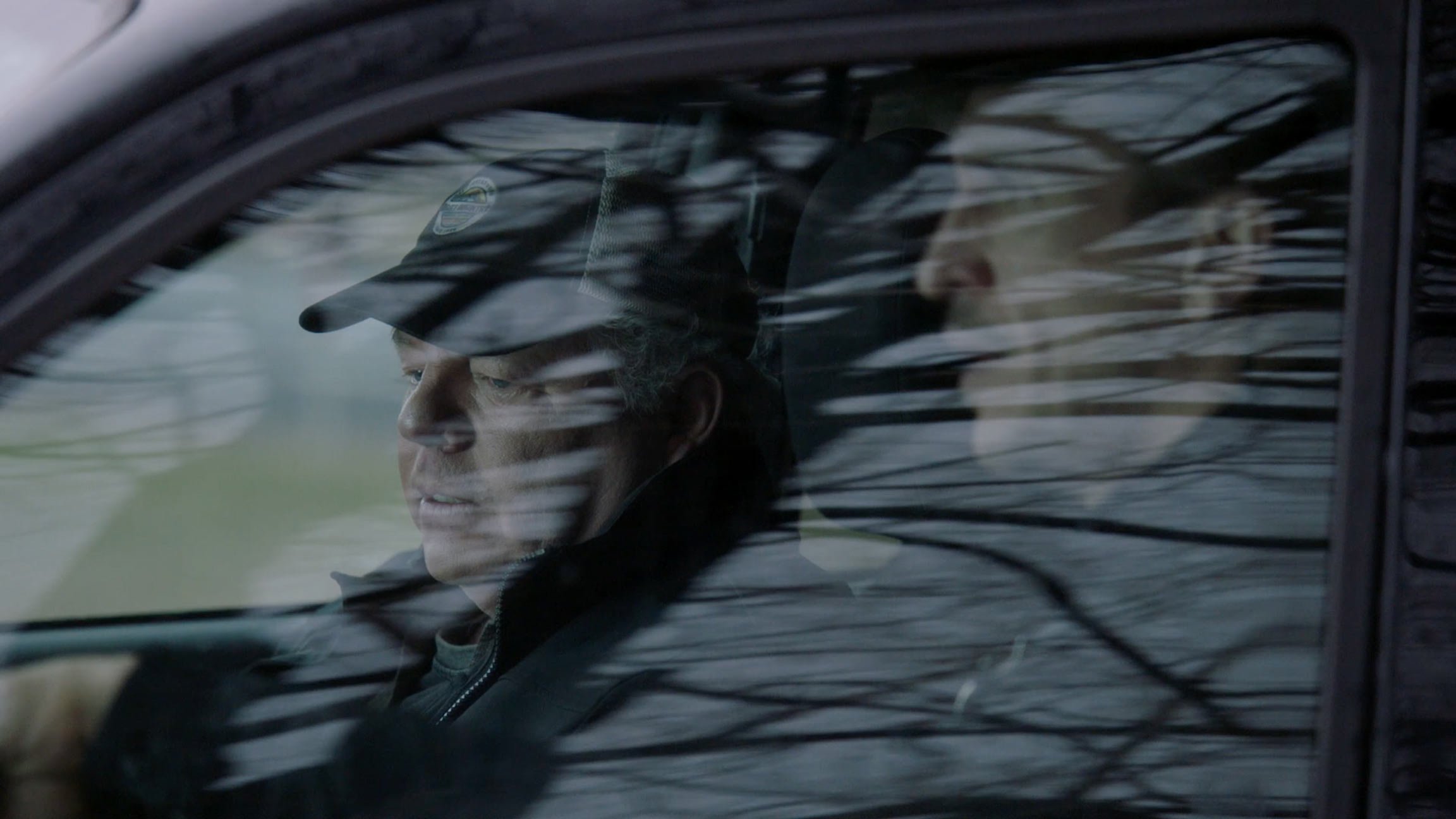 A man with gray hair and a black cap sits in a car, looking downward through the window with reflections of tree branches on the glass.