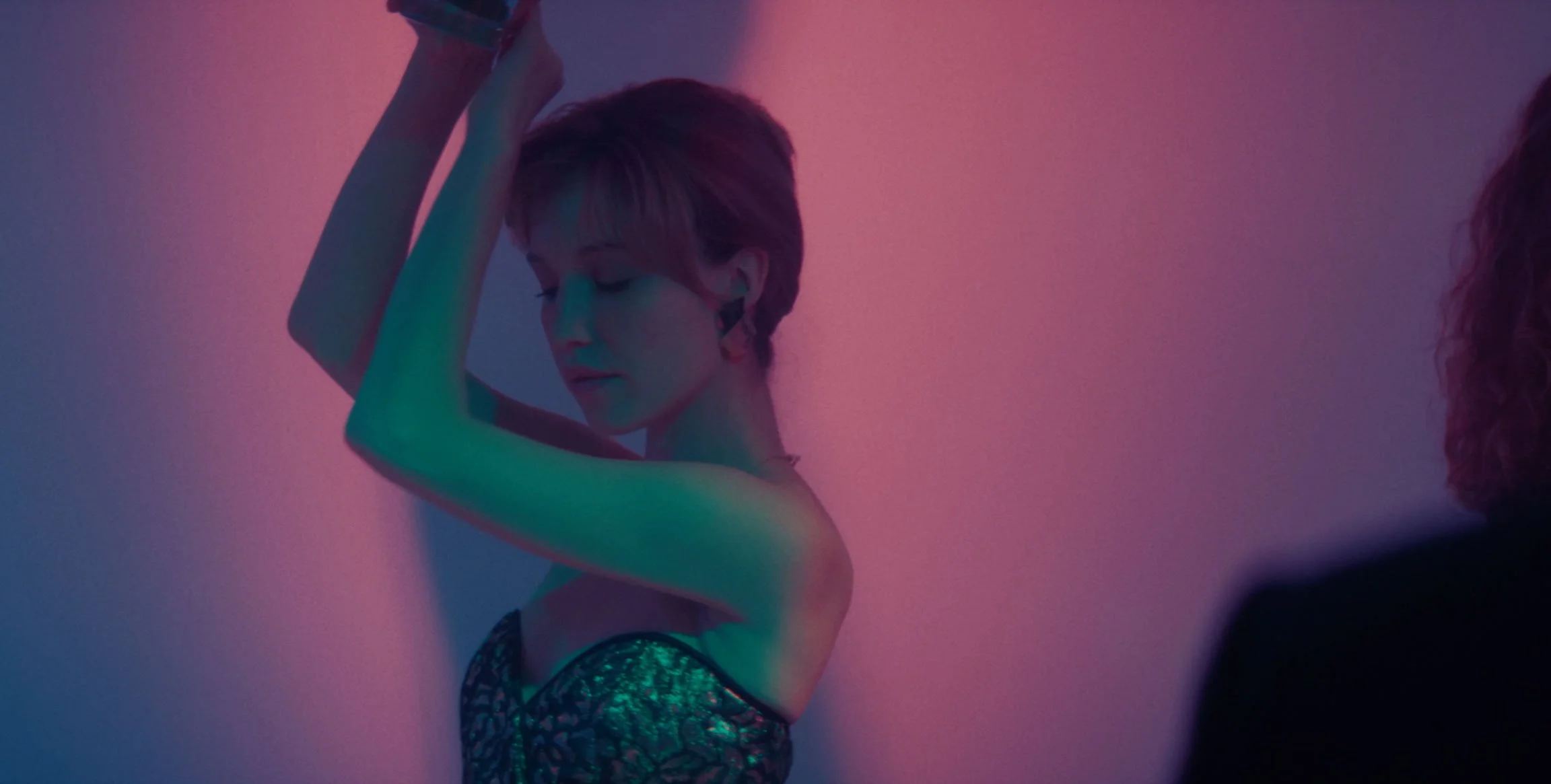 A woman with short hair and a strapless dress is dancing with her eyes closed against a pink and blue gradient background.