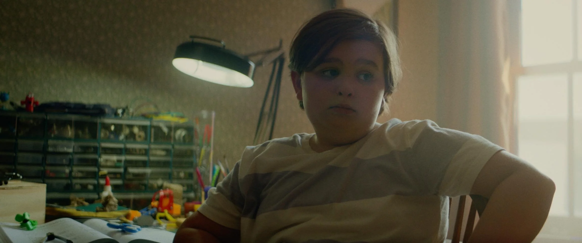A young boy with short brown hair sitting at a desk in a dimly lit room, with a window and curtain in the background. The desk has various toys, a small shelving unit, and an open book.