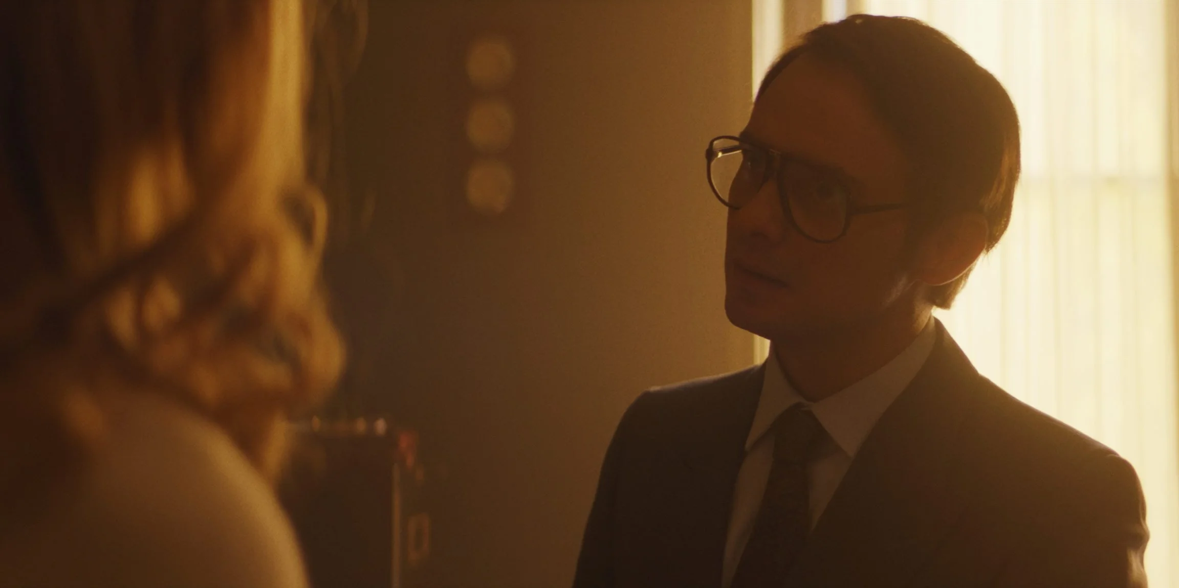 A man with glasses and a suit talking to a woman indoors, backlit by a yellow light from a window, creating a silhouette effect.
