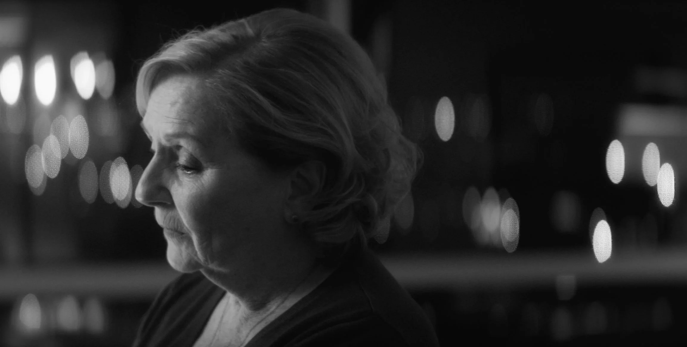 Black and white photo of an elderly woman with her eyes closed, facing left, with her gray hair styled in soft waves, against a background of blurred lights.