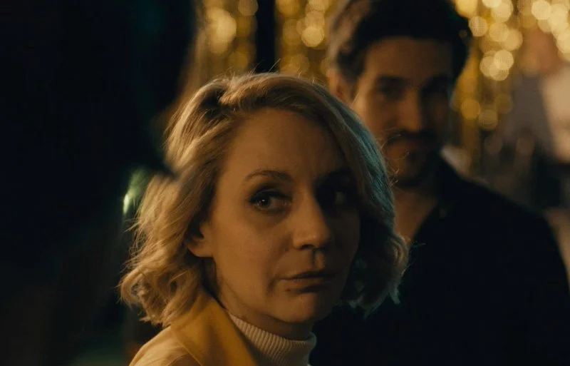 A woman with blonde hair and a yellow jacket looking over her shoulder with a serious expression, in a dimly lit scene with warm lighting.