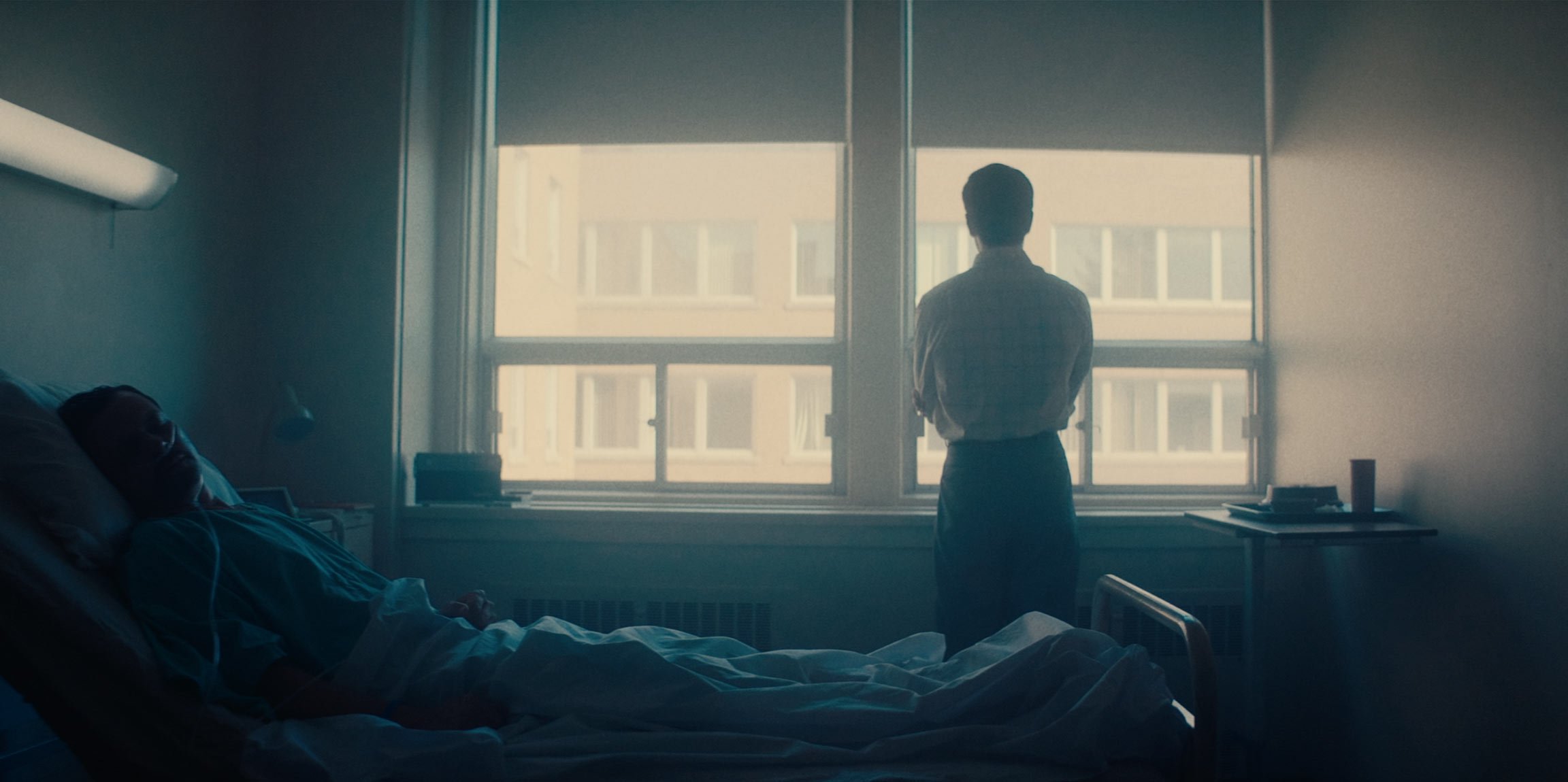 A person in a hospital bed with medical tubes, lying on their side, looking towards a window. Another person stands by the window, gazing outside. The room is softly lit with natural light from the window.