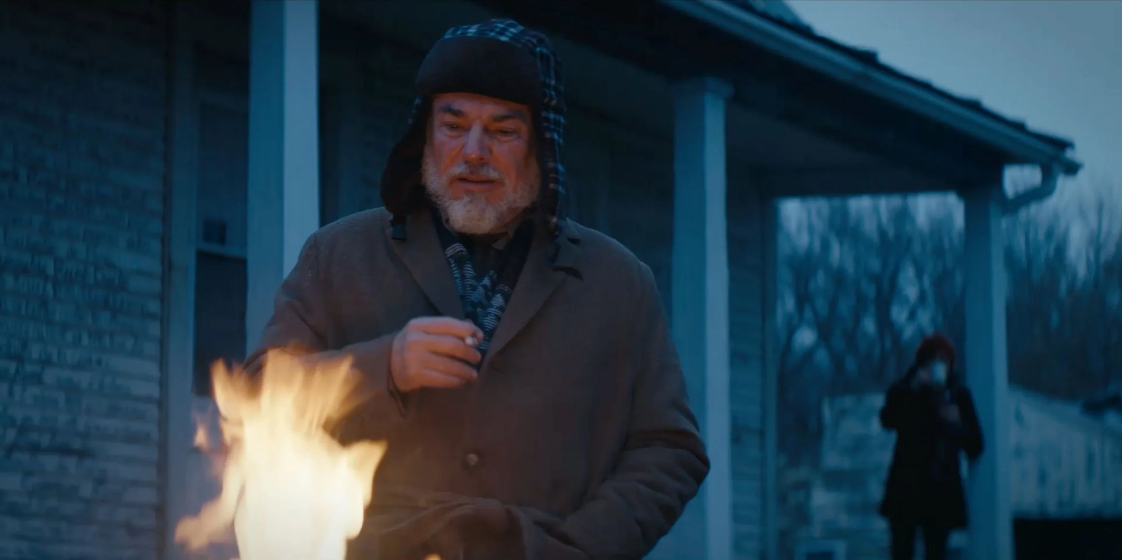 An older man with a beard and a black and blue winter hat standing outside near a small fire, with a woman wearing a mask and dark clothing in the background near a house.