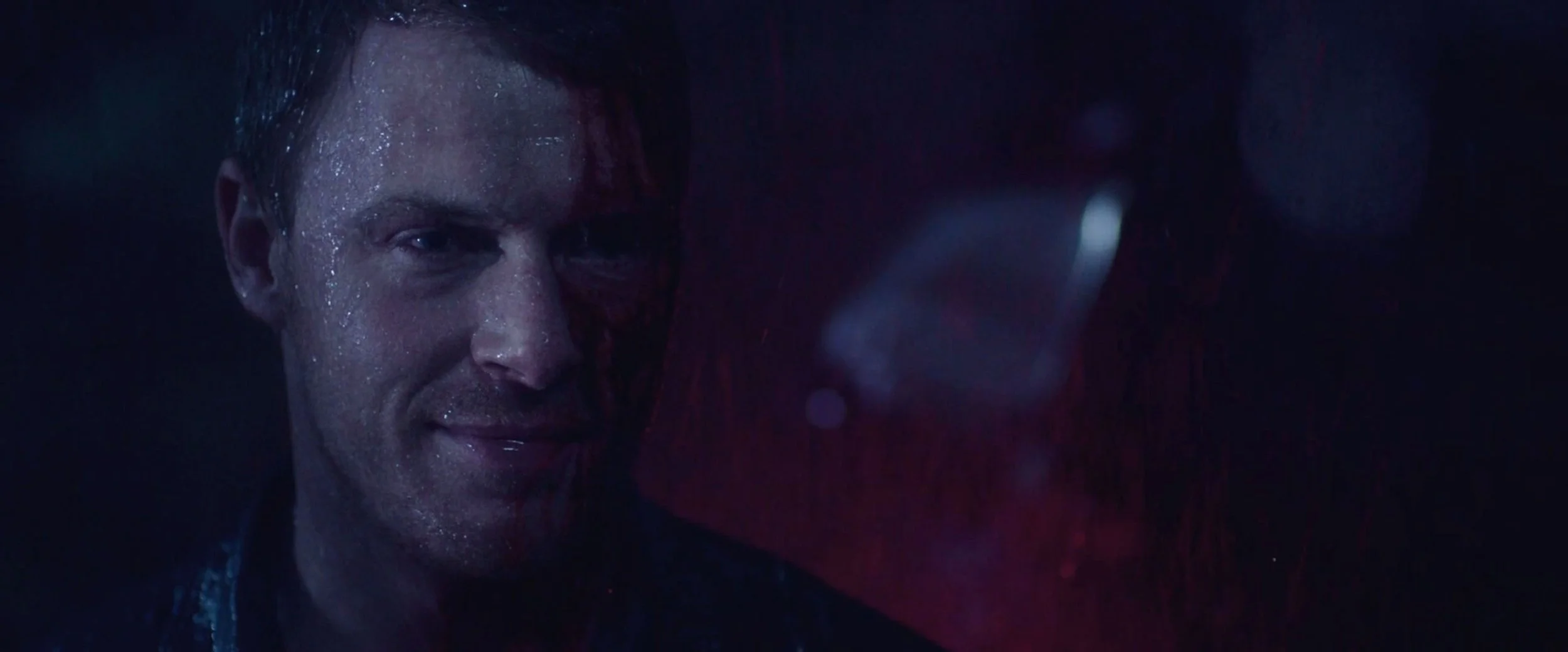 Close-up of a man smiling with wet face in a dark setting, with a blurred red object nearby.