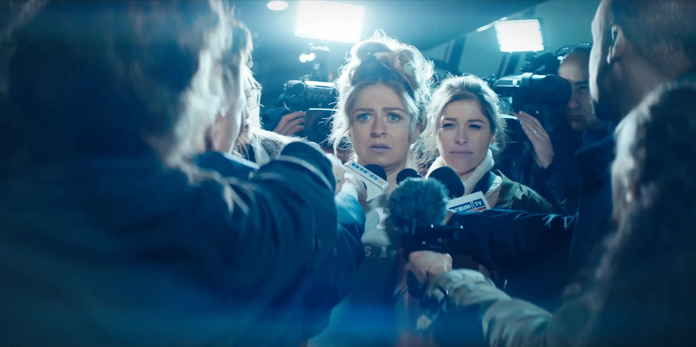 A woman with blonde hair surrounded by reporters and microphones during a press conference or interview.
