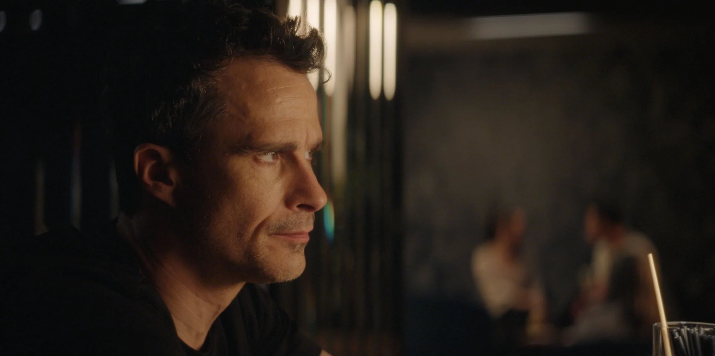 A man with short dark hair staring thoughtfully in a dimly lit setting, with blurred people in the background.