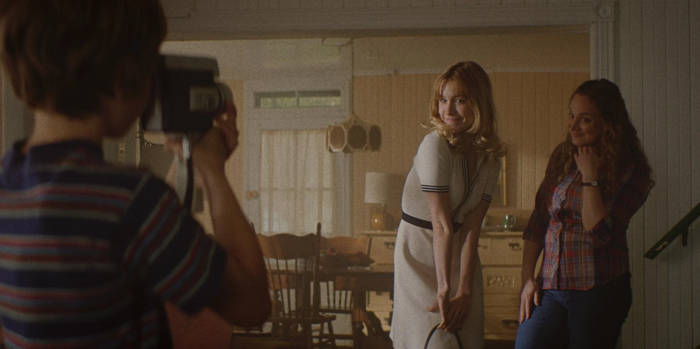 A young girl with short hair taking a photo of two women in a cozy, vintage-style kitchen. One woman has red hair, wearing a white dress with black stripes, and is smiling while posing. The other woman has curly hair, wearing a plaid shirt, and is sm