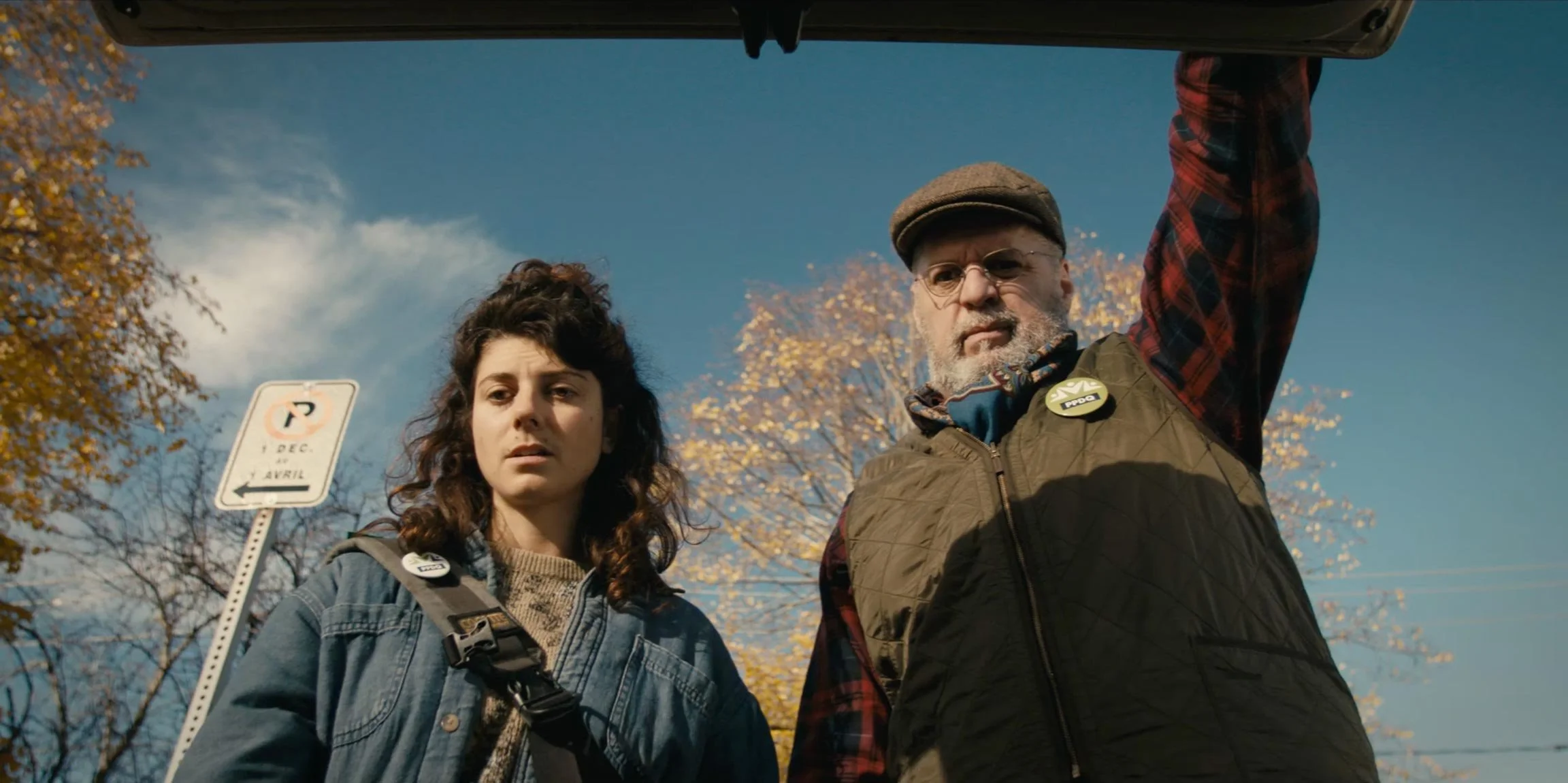 A young woman and an older man are looking down at the camera from underneath a vehicle's open hood, with a stop sign and autumn trees with yellow leaves in the background.