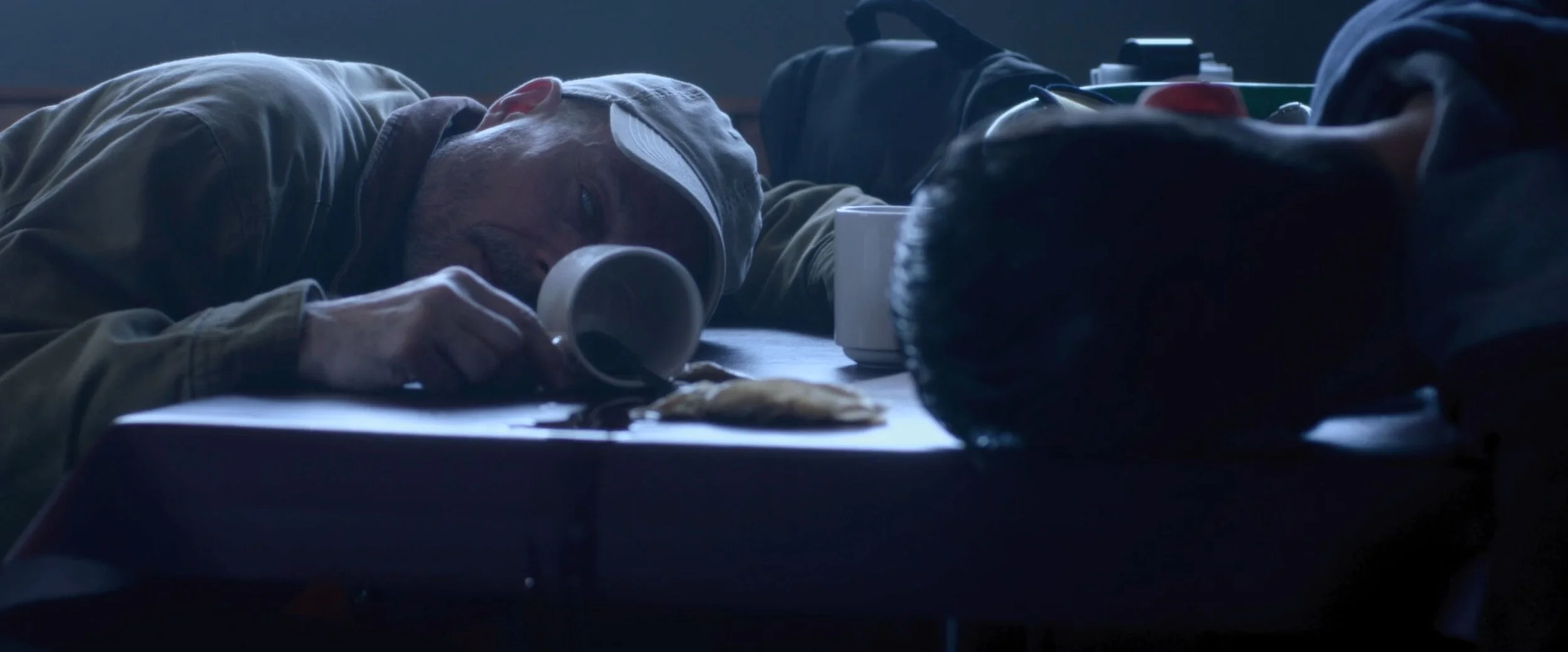 A man lying on a table, resting his head on his arm, drinking from a mug with a biscuit and crumbs nearby.