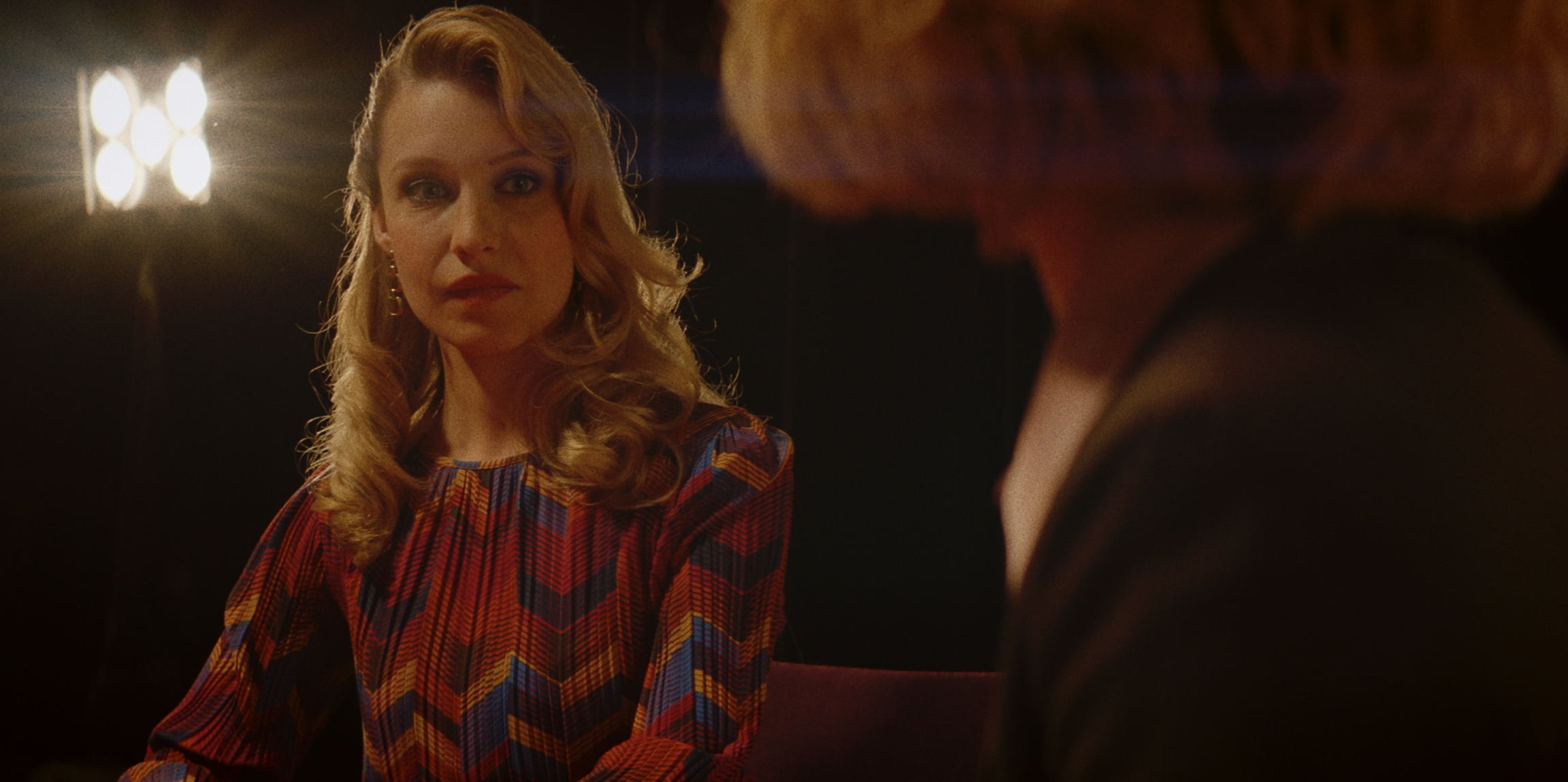 A woman with red hair and a colorful patterned dress looks at a person with blonde hair in a dark room with a bright light in the background.