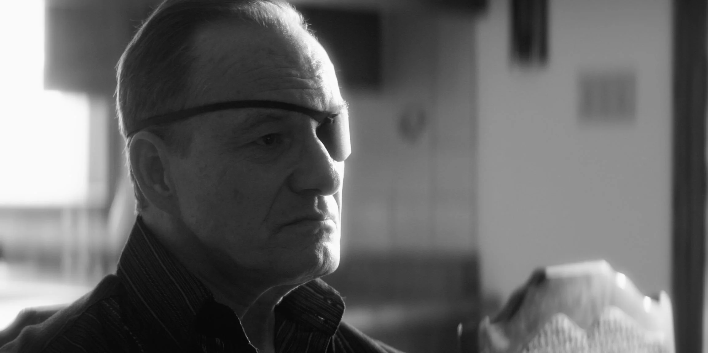 Black and white close-up of a man with short hair, wearing an eye patch over his right eye, sitting indoors with a serious expression.