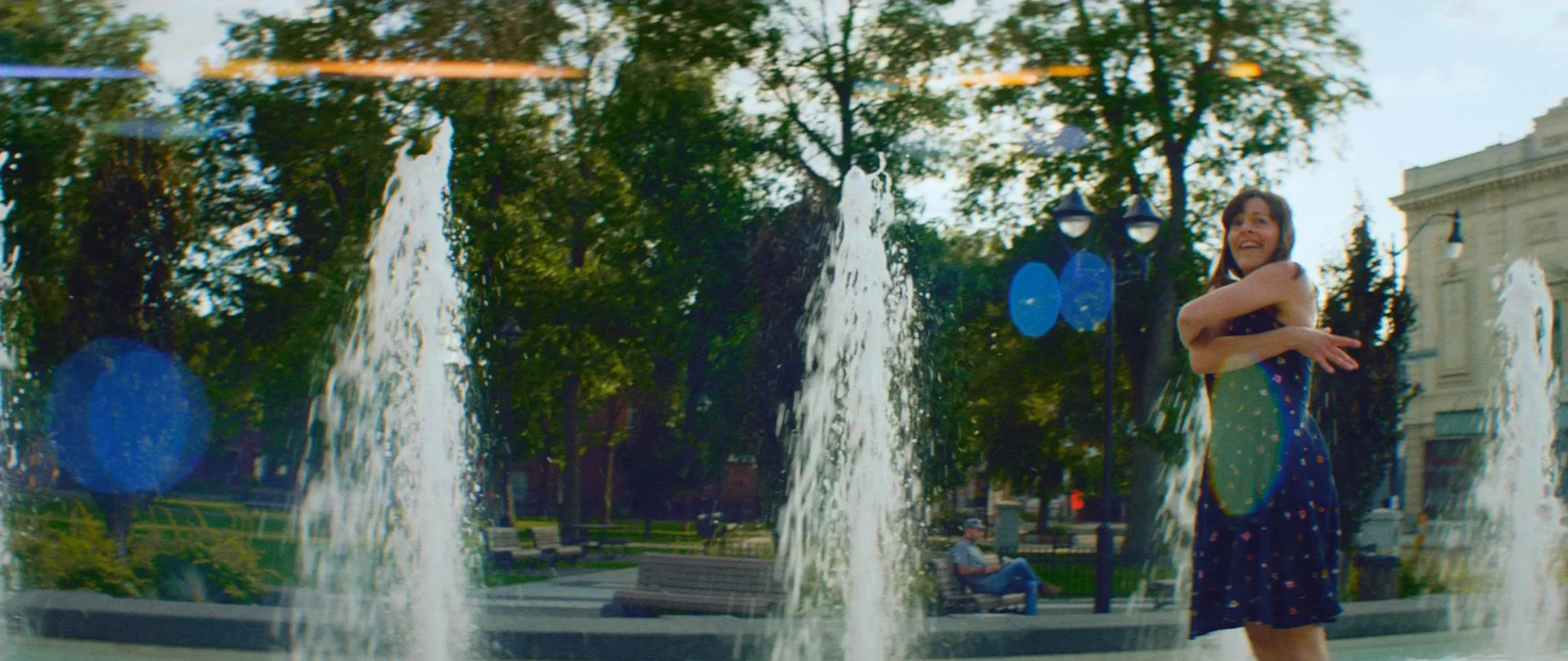 A joyful woman in a blue dress with yellow patterns standing by a fountain in a park, surrounded by trees, benches, and street lamps, with a person sitting on a bench in the background.